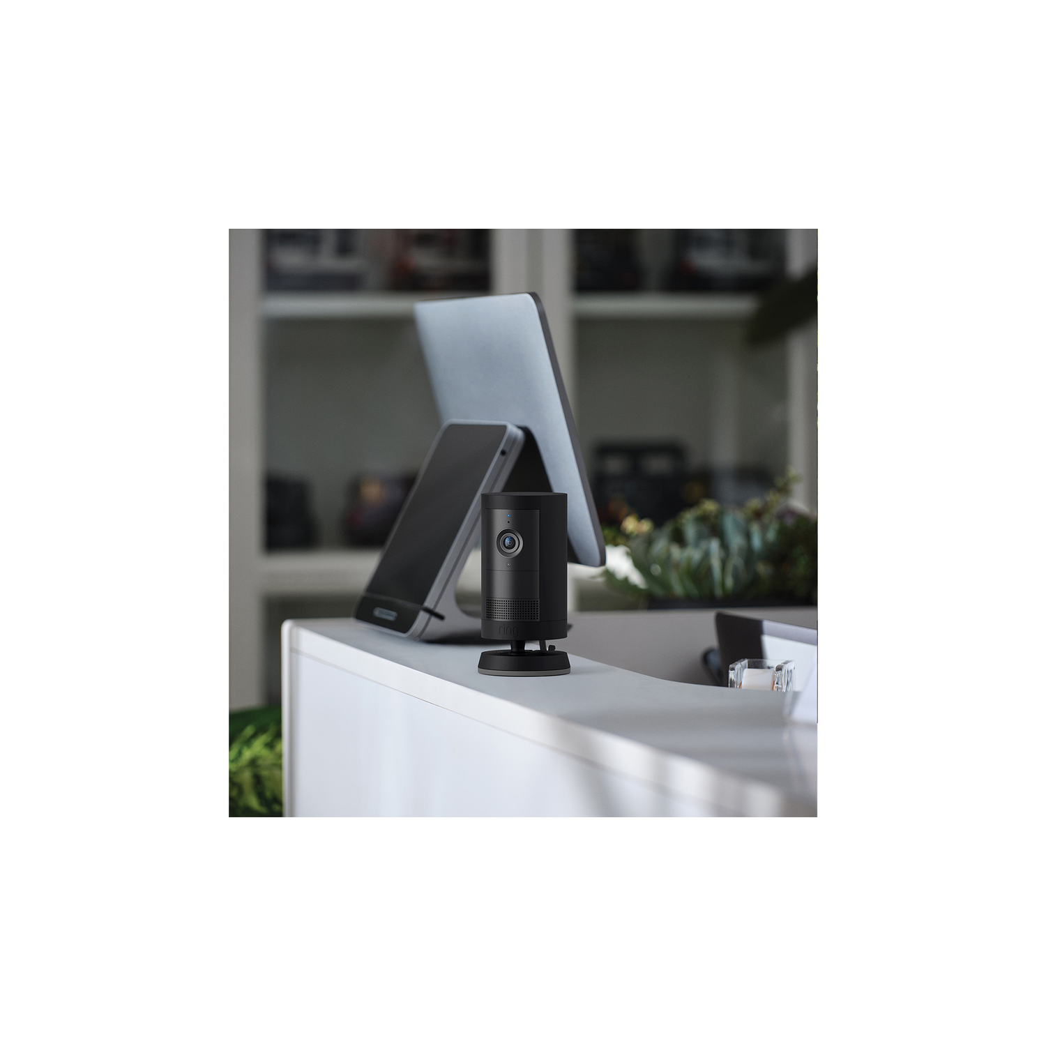 A black Ring camera sits on a white desk. A laptop and phone are in the background.