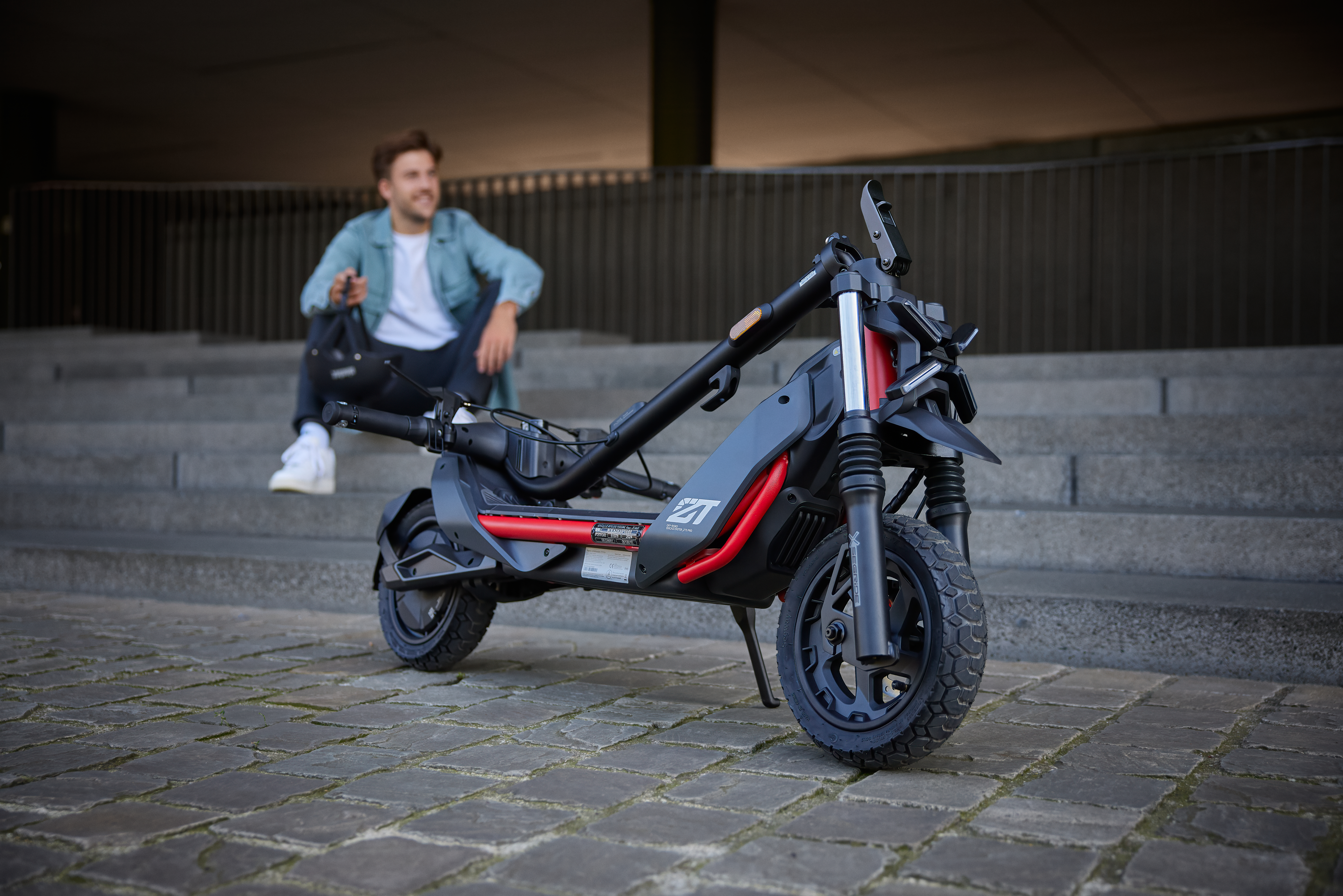 Bir adam gülümsüyor, basamakların yanında oturuyor, e-scooter kısmen katlanmış, kırmızı ve siyah.