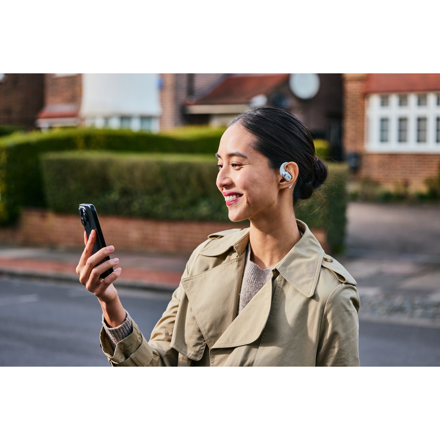 Woman holding phone, wearing earbuds, smiling, outside.