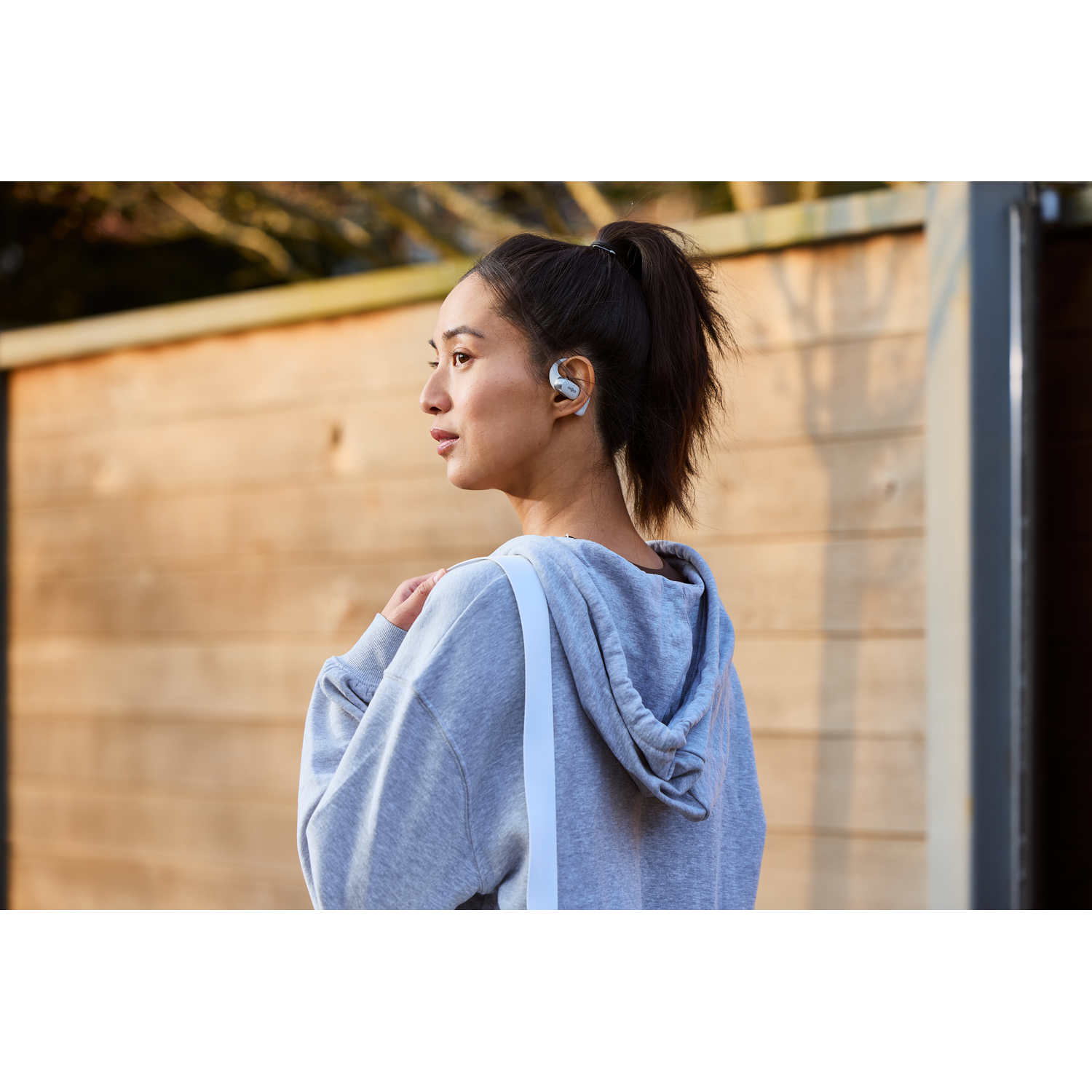 Woman wearing earbuds, looking away, in front of wooden fence.