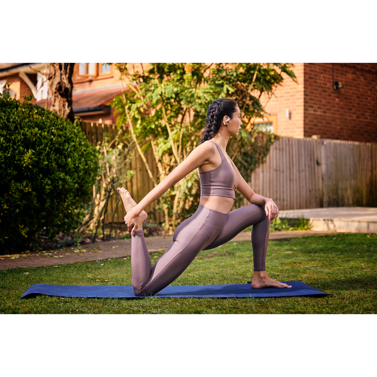 Woman doing yoga, wearing earbuds, on mat, outside. Green background.