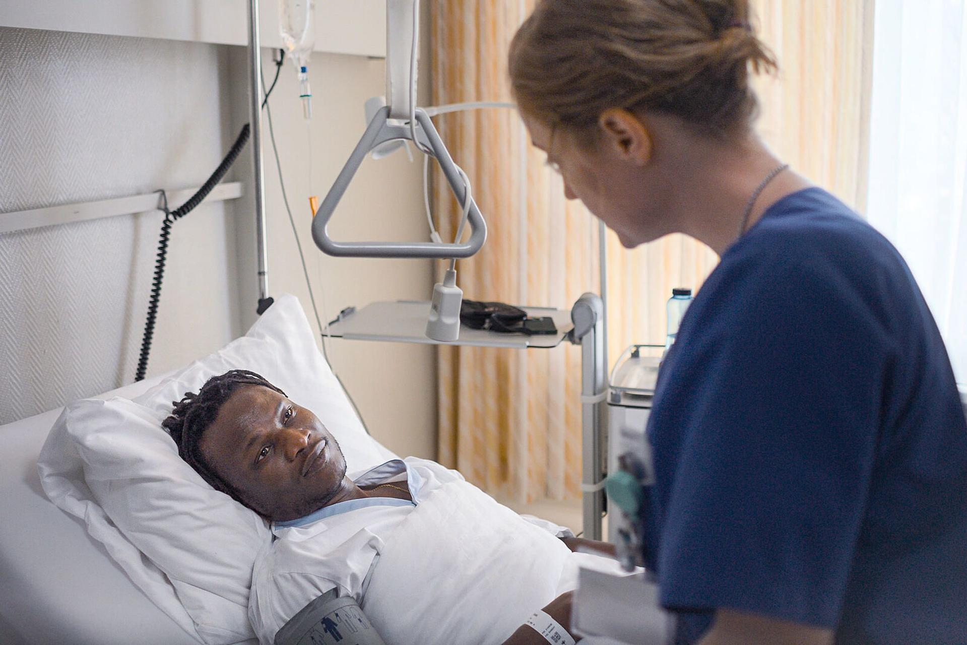 Krankenschwester in blauer OP-Kleidung spricht mit einem Patienten im Bett.