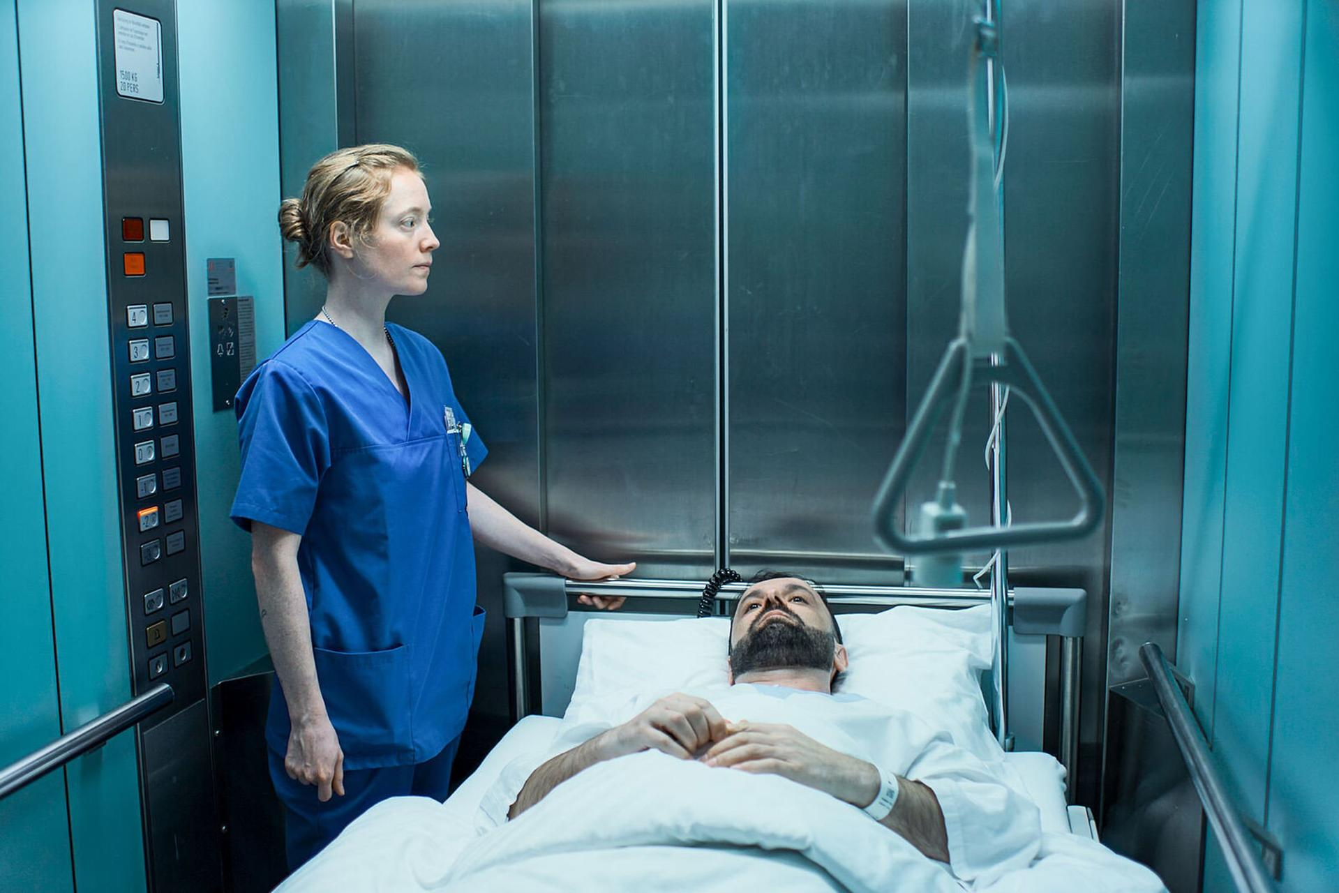 Eine Krankenschwester und ein Patient in einem Aufzug. Der Patient liegt im Bett und schaut nach oben.