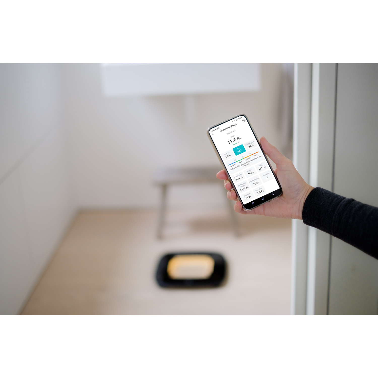 Persoon die een smartphone vasthoudt met gezondheidsappgegevens. Weegschaal op de achtergrond zichtbaar.