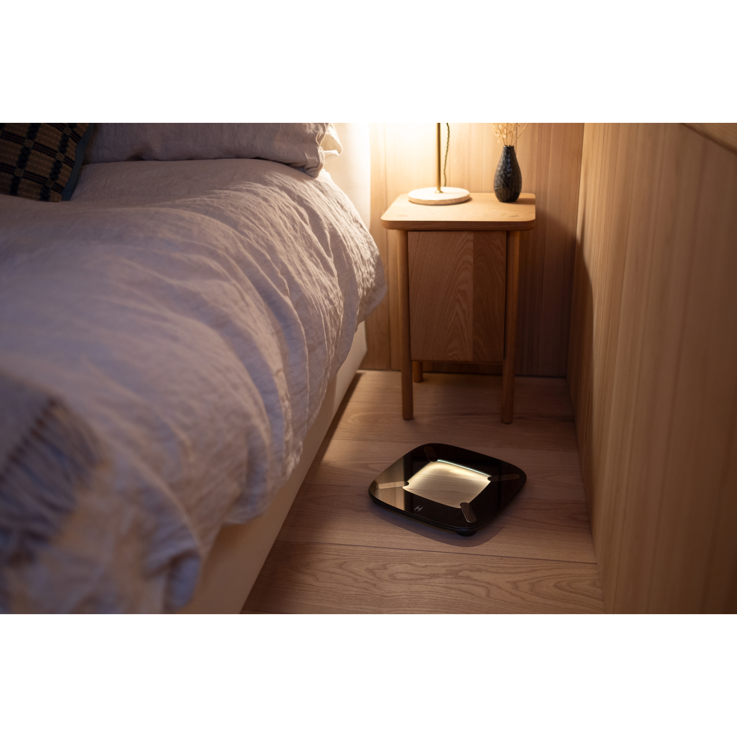 Slimme weegschaal op houten vloer naast een bed, een lamp en een nachtkastje. Zachte verlichting.