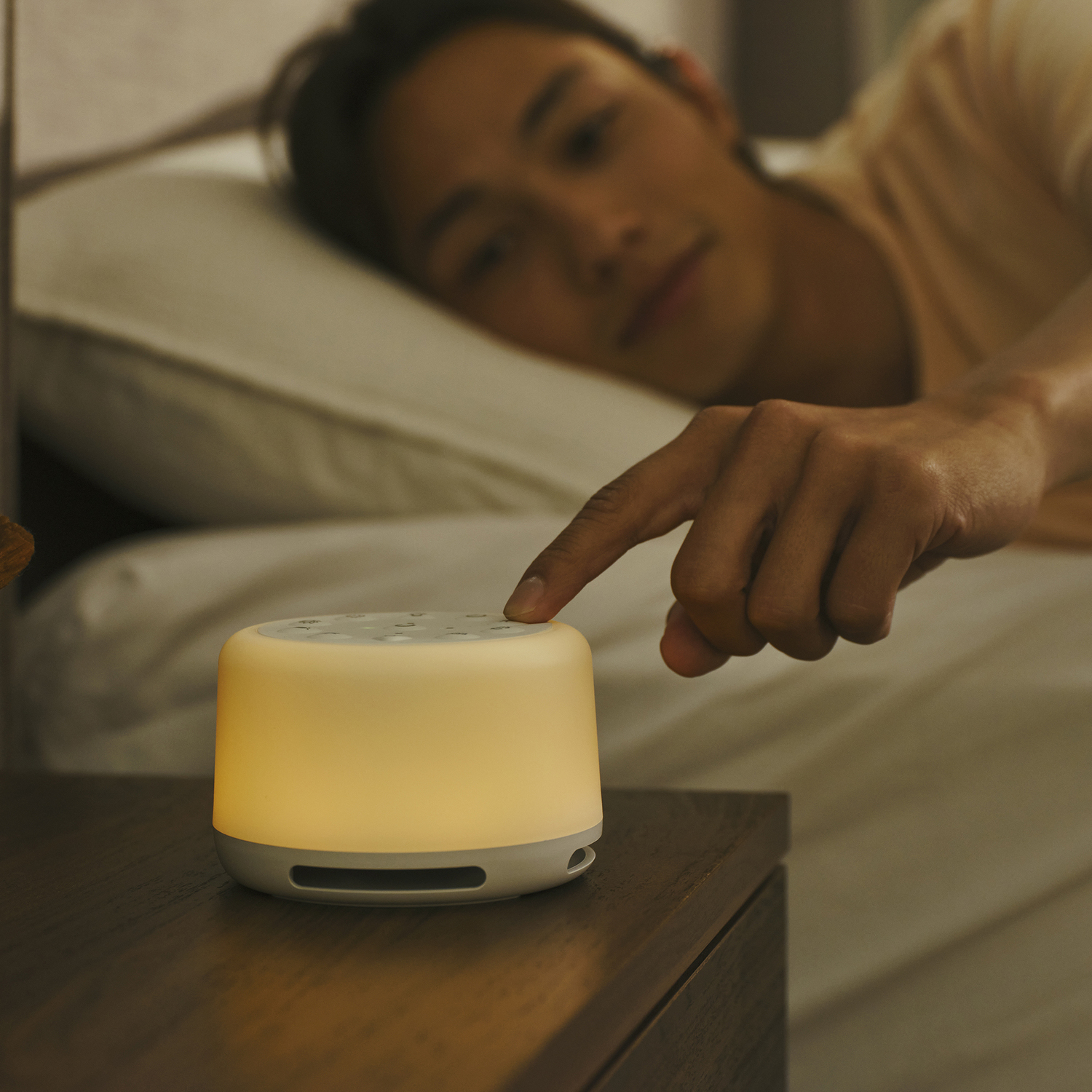 Man in bed, reaching to turn on a sound machine. Warm light, on a nightstand.