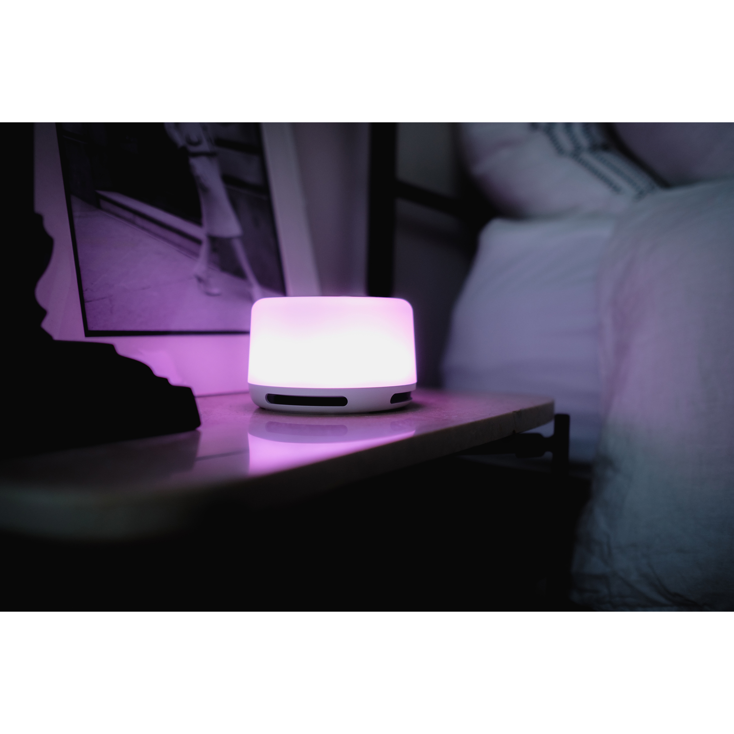 Sound machine on nightstand emitting purple light. Beside a bed, lamp, and framed picture.