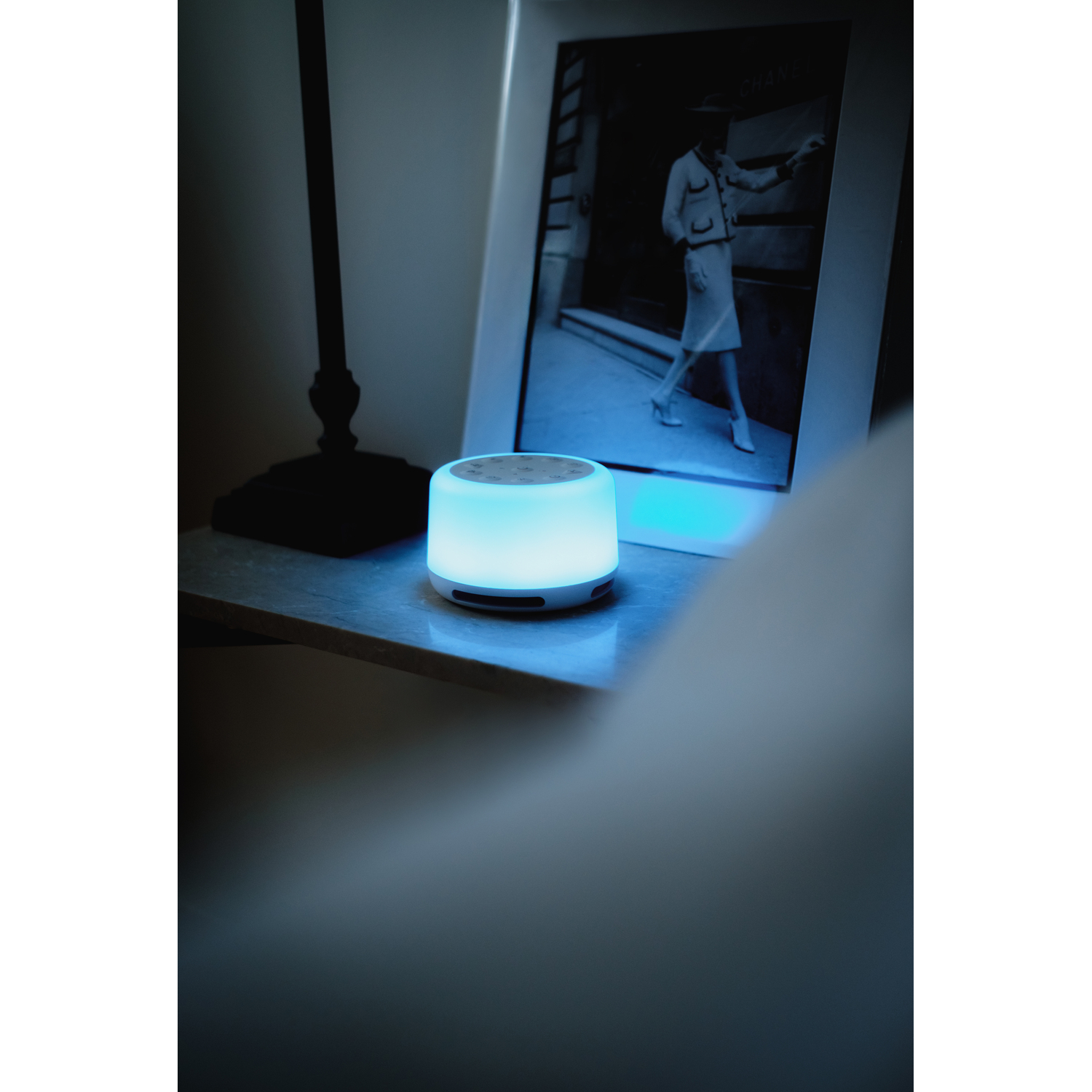 Sound machine on nightstand emitting blue light. Beside a bed, lamp, and framed picture.