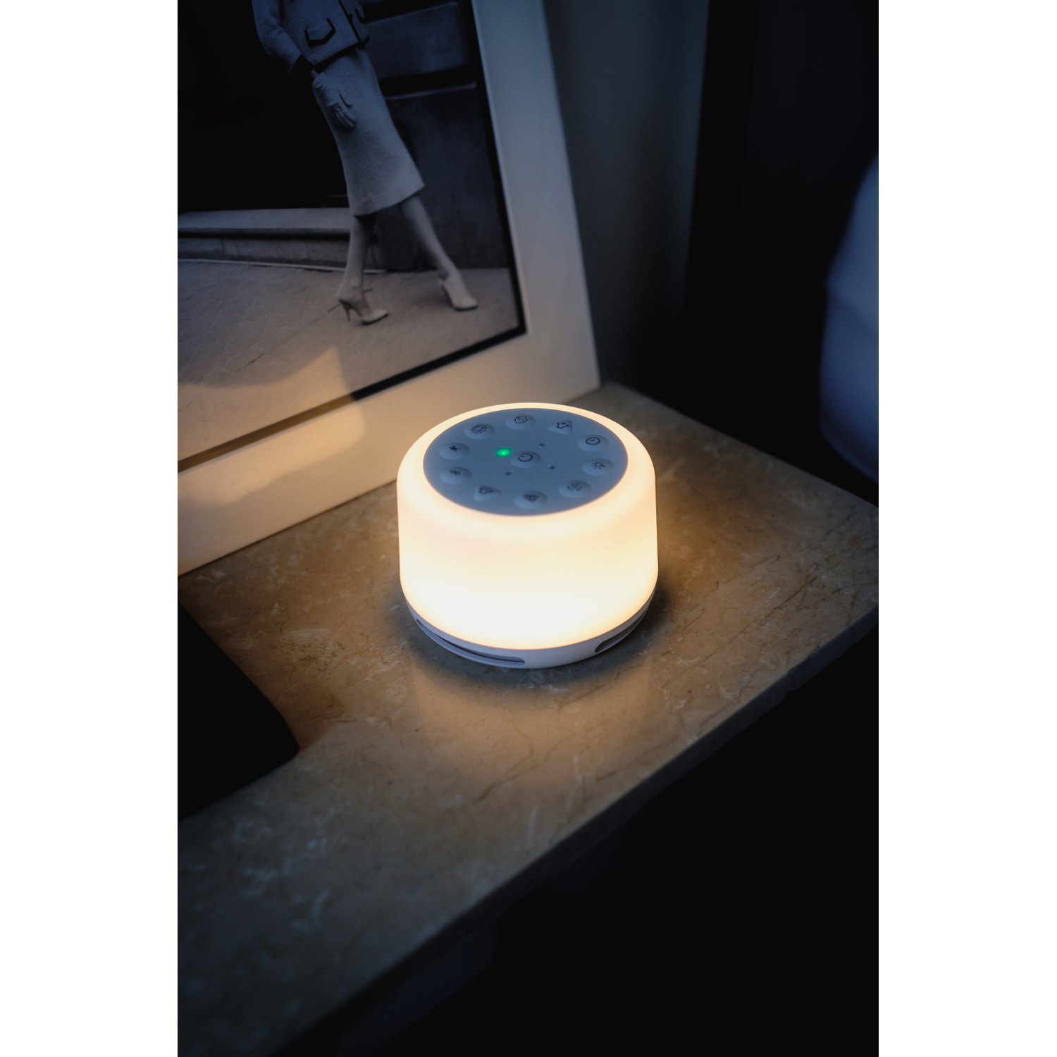 Glowing white and yellow sound machine on a table, with a framed black and white image in the background.