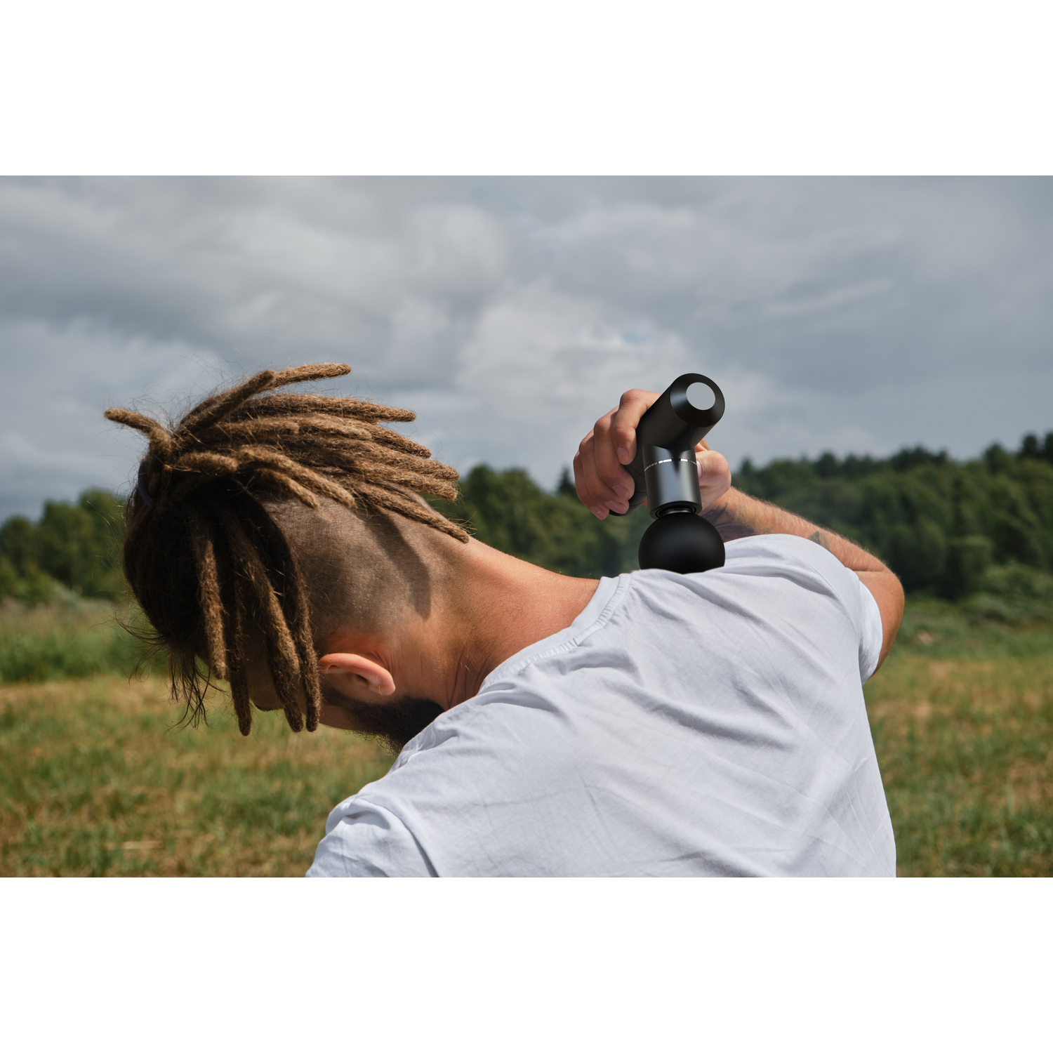 A man with dreadlocks uses a massage gun on his neck, outdoors with a cloudy sky.