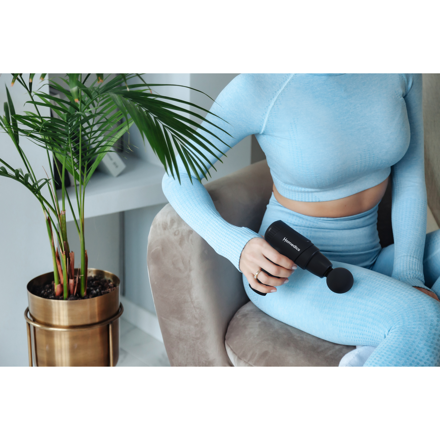 Woman in blue athletic wear holds a black massage gun near a houseplant and a chair.