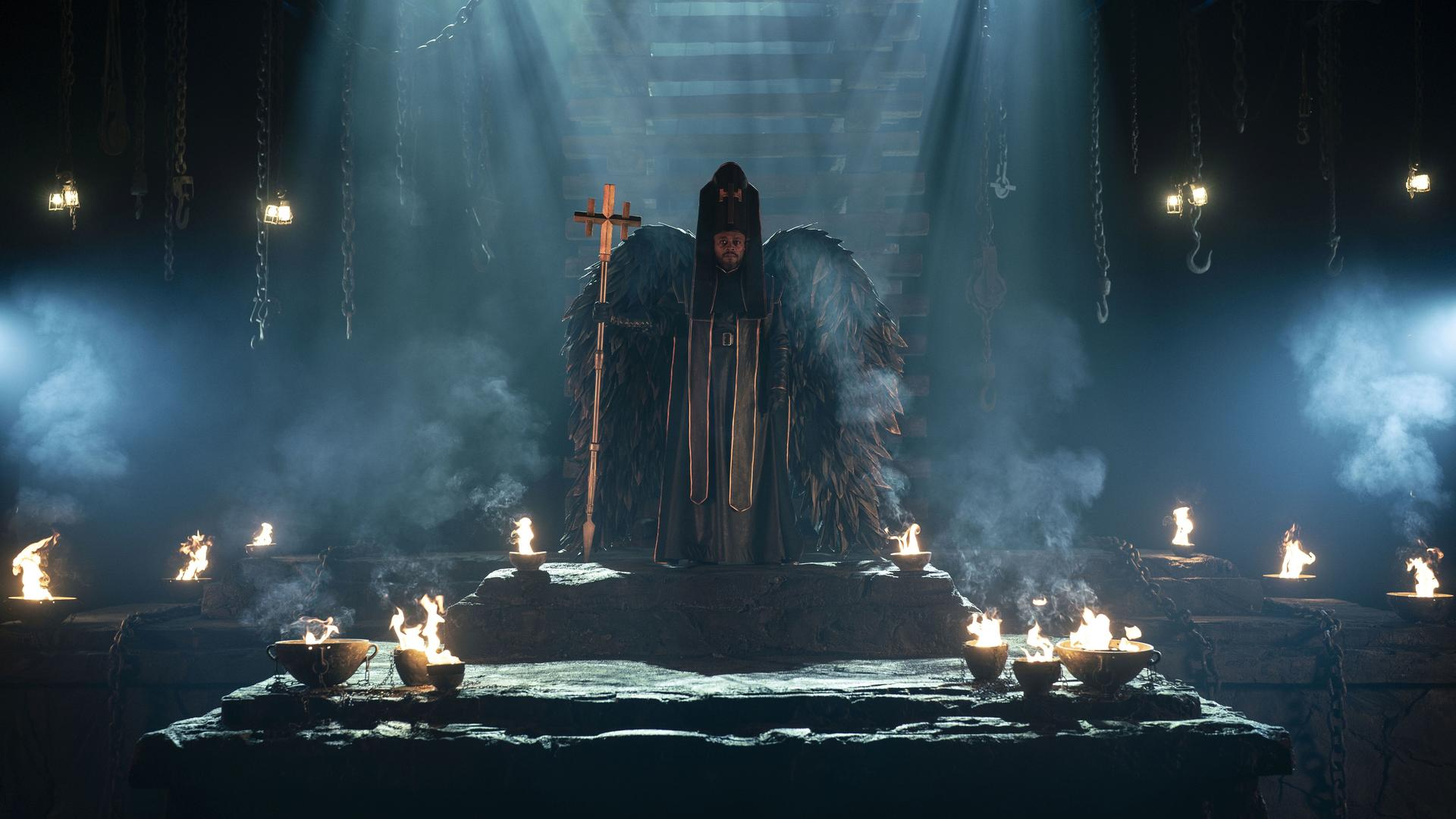 Figur in dunklen Roben, Flügel, hält Kreuz. Beleuchtet von Kerzen, in dunkler Umgebung.