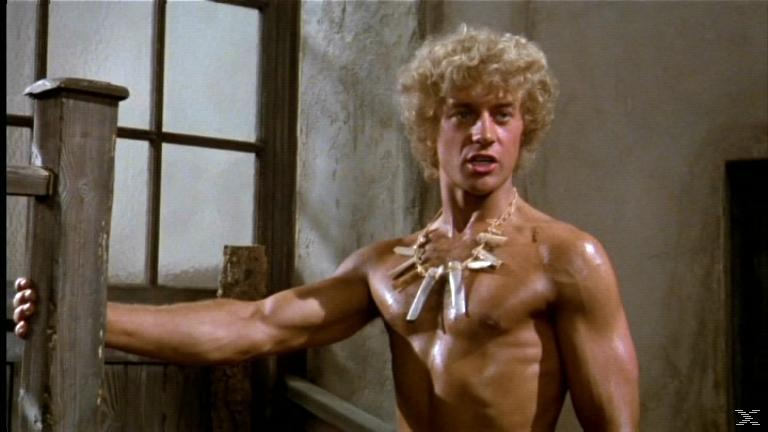 Ein Mann ohne Hemd mit blonden Locken lehnt an einem Holzpfosten.