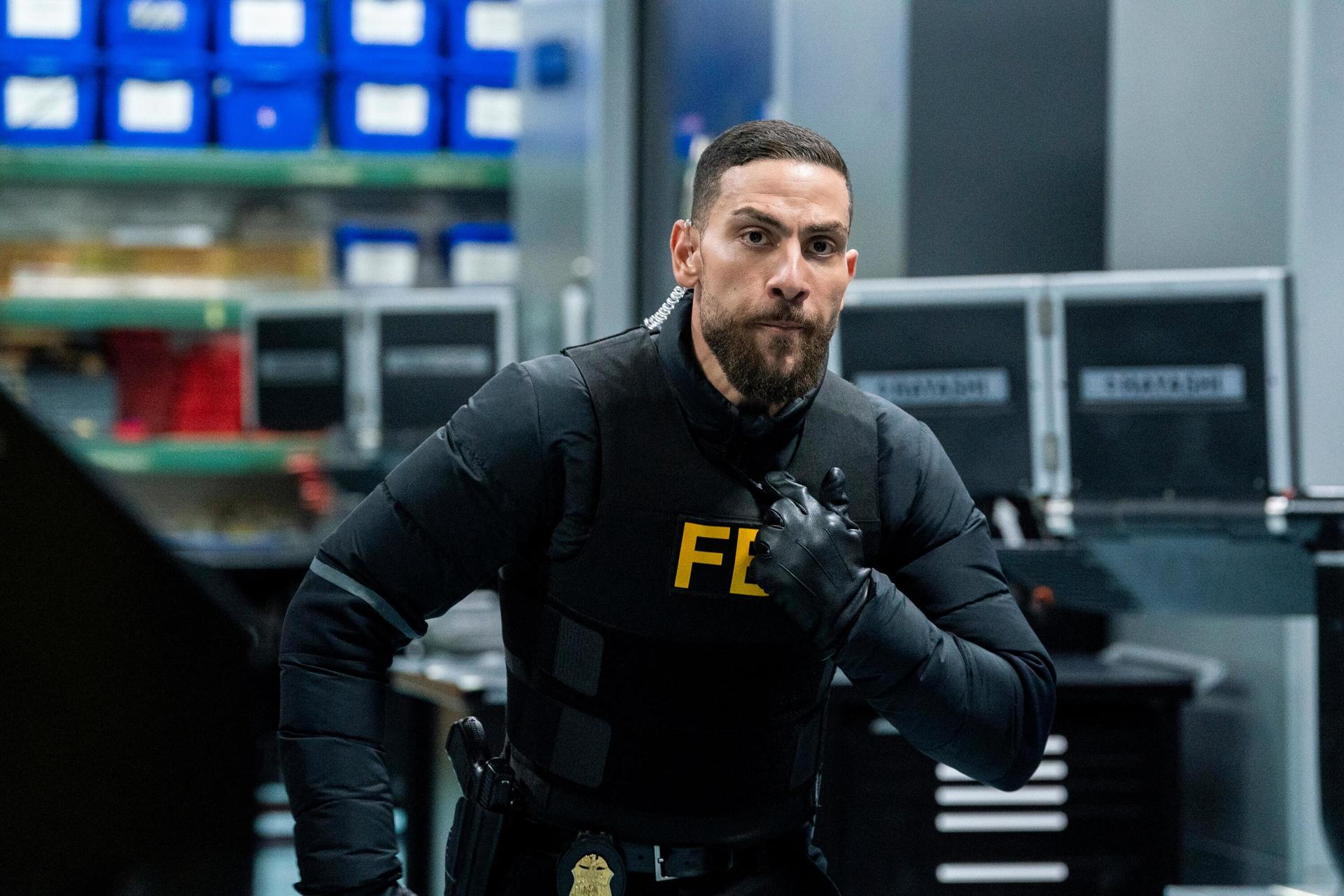 Ein FBI-Agent, der eine Weste, Handschuhe und ein Headset trägt, blickt aufmerksam. Im Hintergrund blaue Behälter.