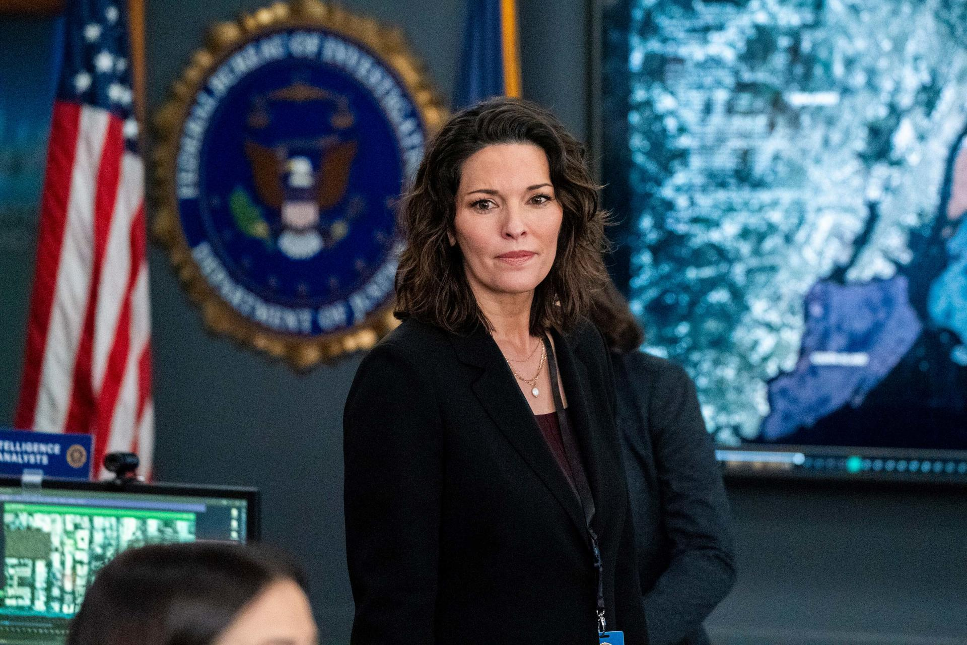 Frau in schwarzem Blazer steht in einem Büro. US-Flagge und FBI-Siegel sind im Hintergrund.