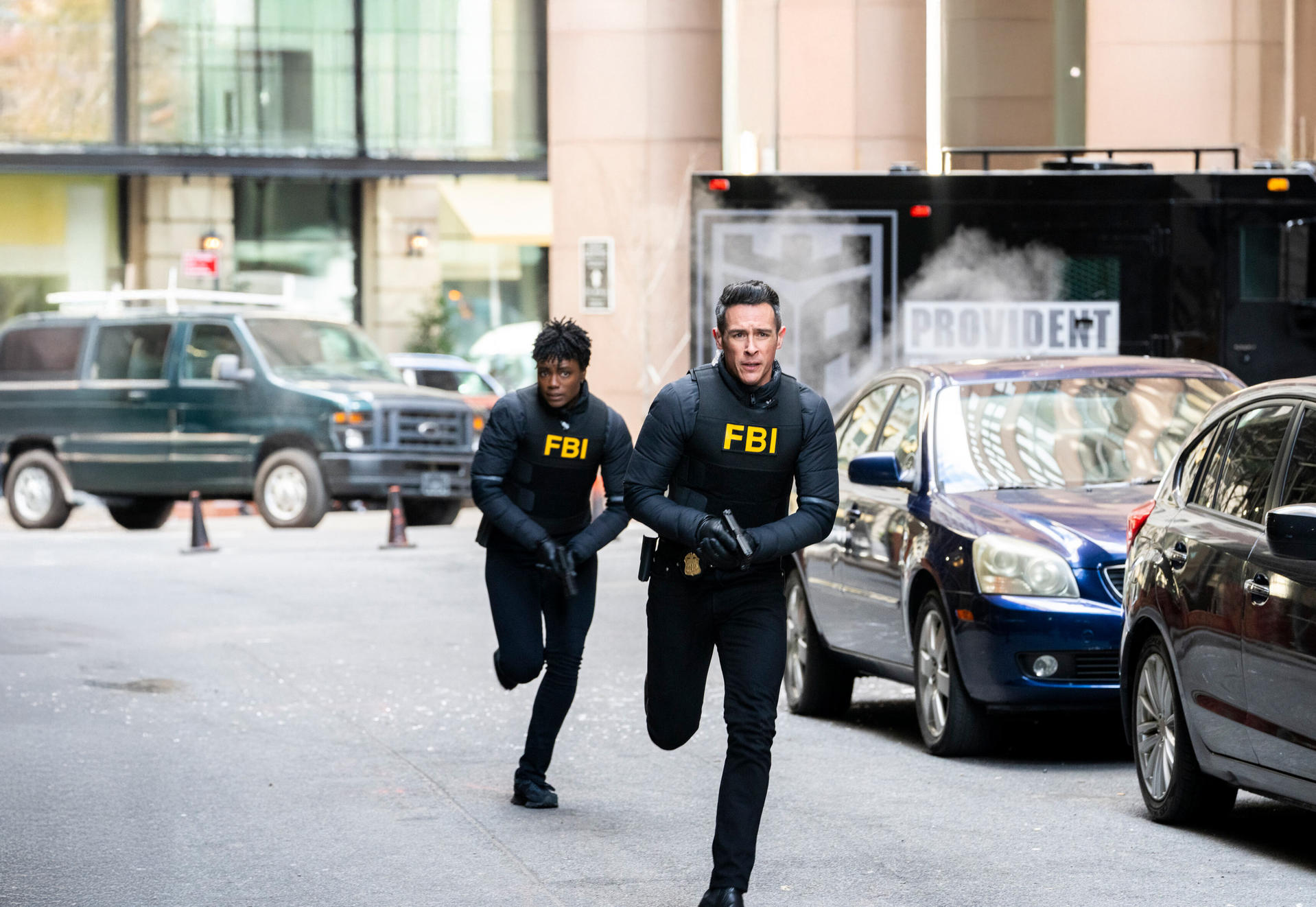 Zwei FBI-Agenten rennen eine Straße entlang, einer hält eine Waffe. Autos und ein Transporter im Hintergrund.