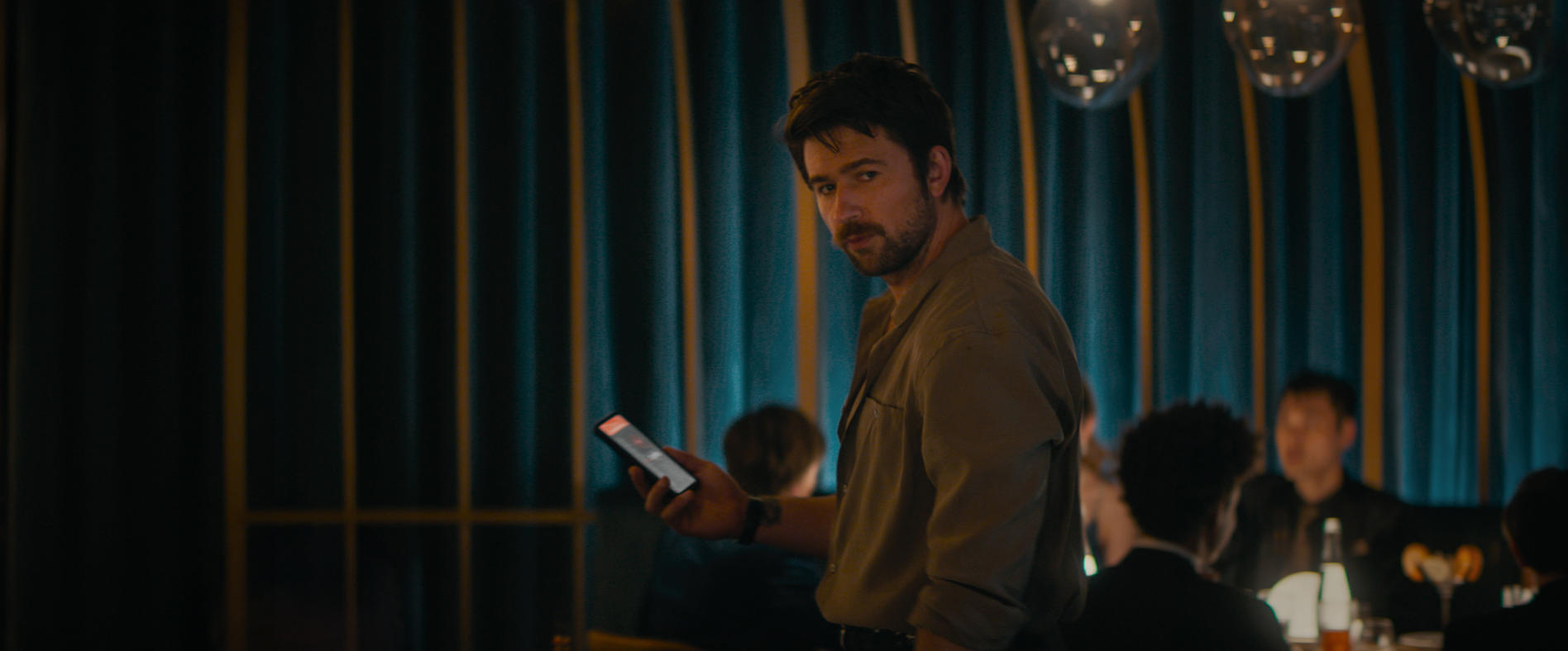 Ein Mann im Hemd hält ein Telefon in einer Bar. Menschen und Lichter im Hintergrund.