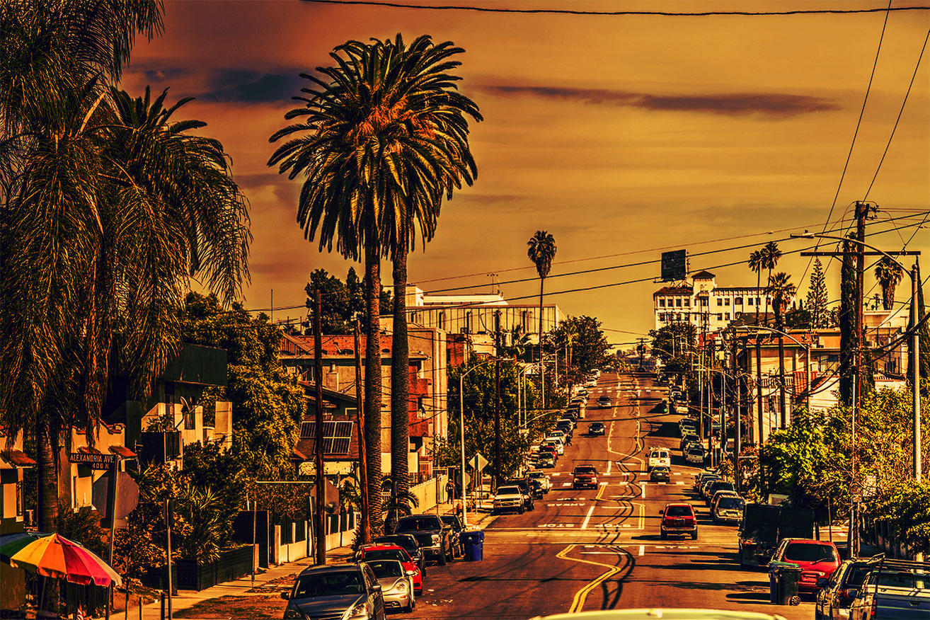 Eine Straße in LA mit Palmen, Autos und Gebäuden unter einem orangefarbenen Himmel.