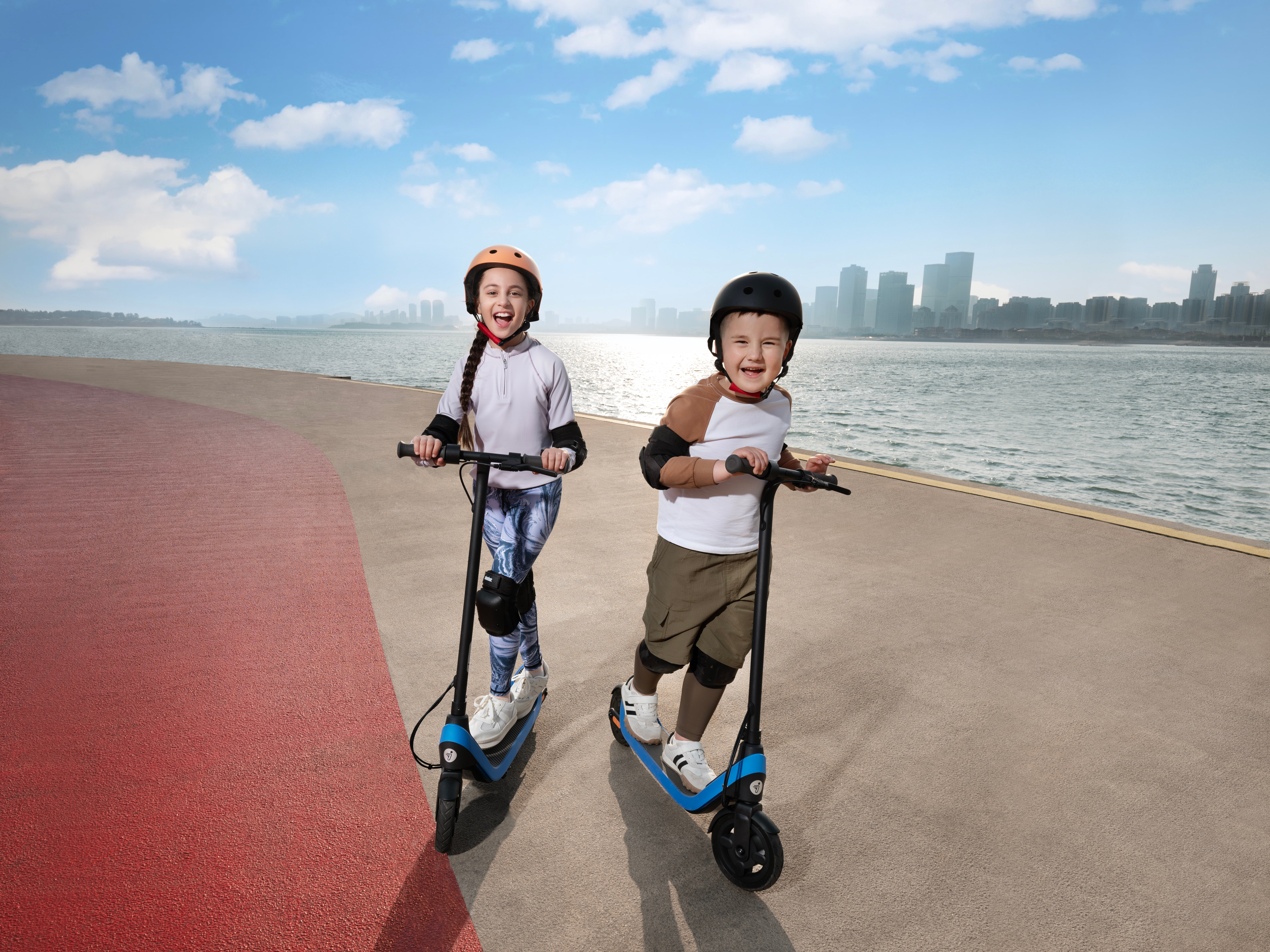 İki çocuk patika üzerinde scooter sürüyor. Gülümsüyorlar ve kask ve dizlik takıyorlar.