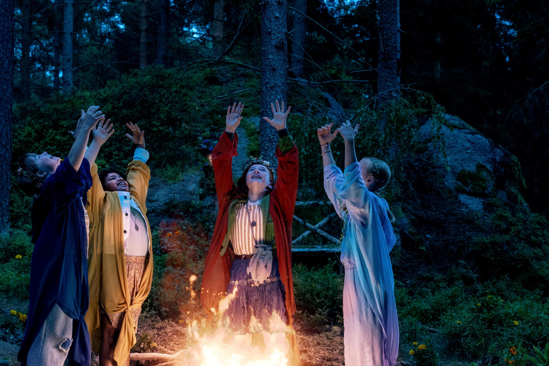 Vier Kinder um ein Feuer, heben die Hände. Sie sind nachts in einem dunklen Wald.