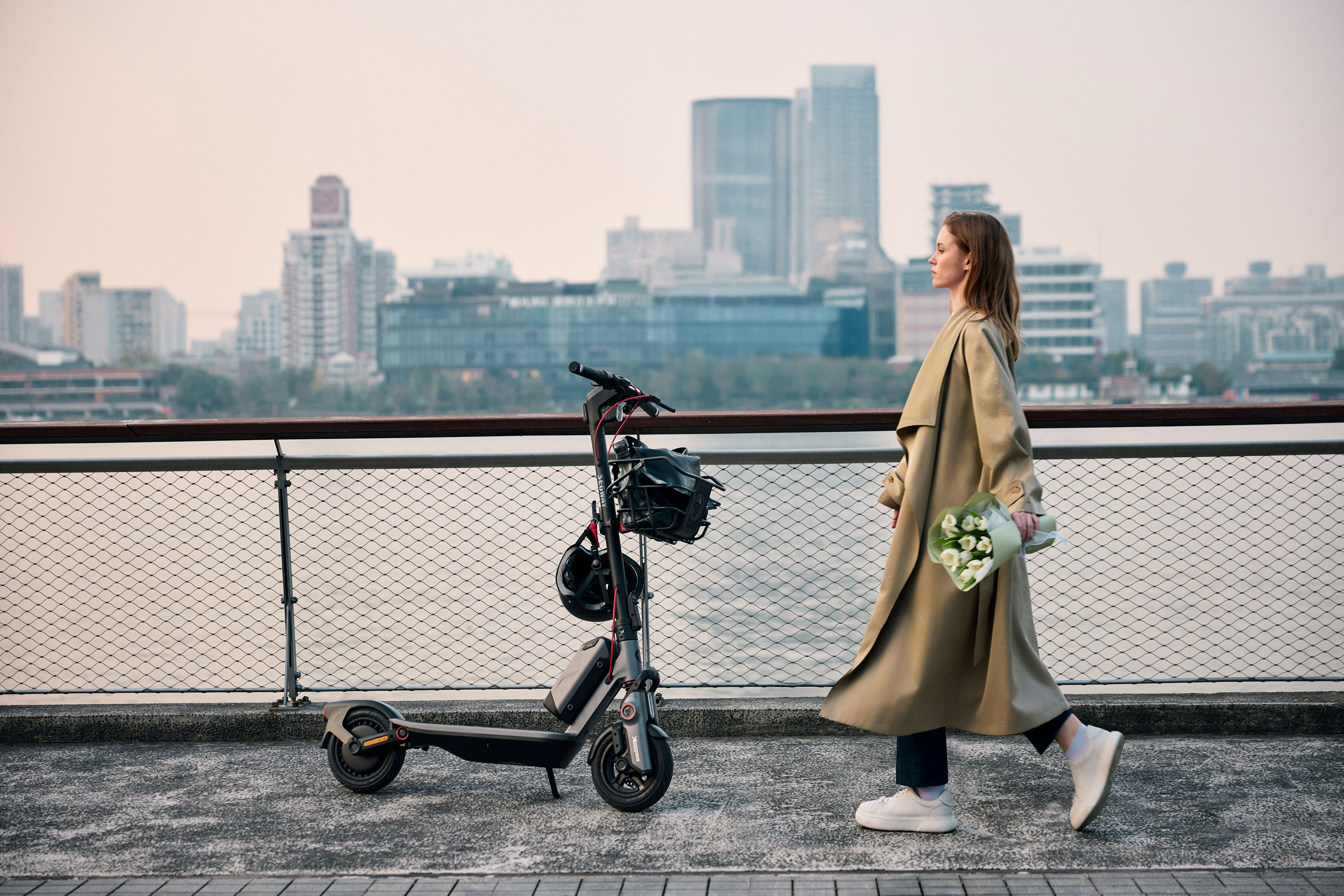 Bir kadın, elinde çiçeklerle bir scooter ve şehir binalarının yanından yürüyor. Gündüz vakti.