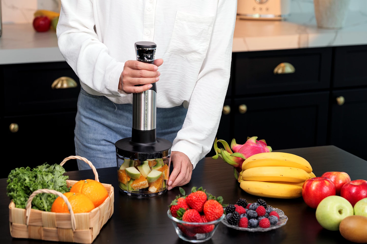 Person hält einen Mixer, der Früchte und Gemüse in einem transparenten Behälter mixt, mit Früchten auf dem Tisch.