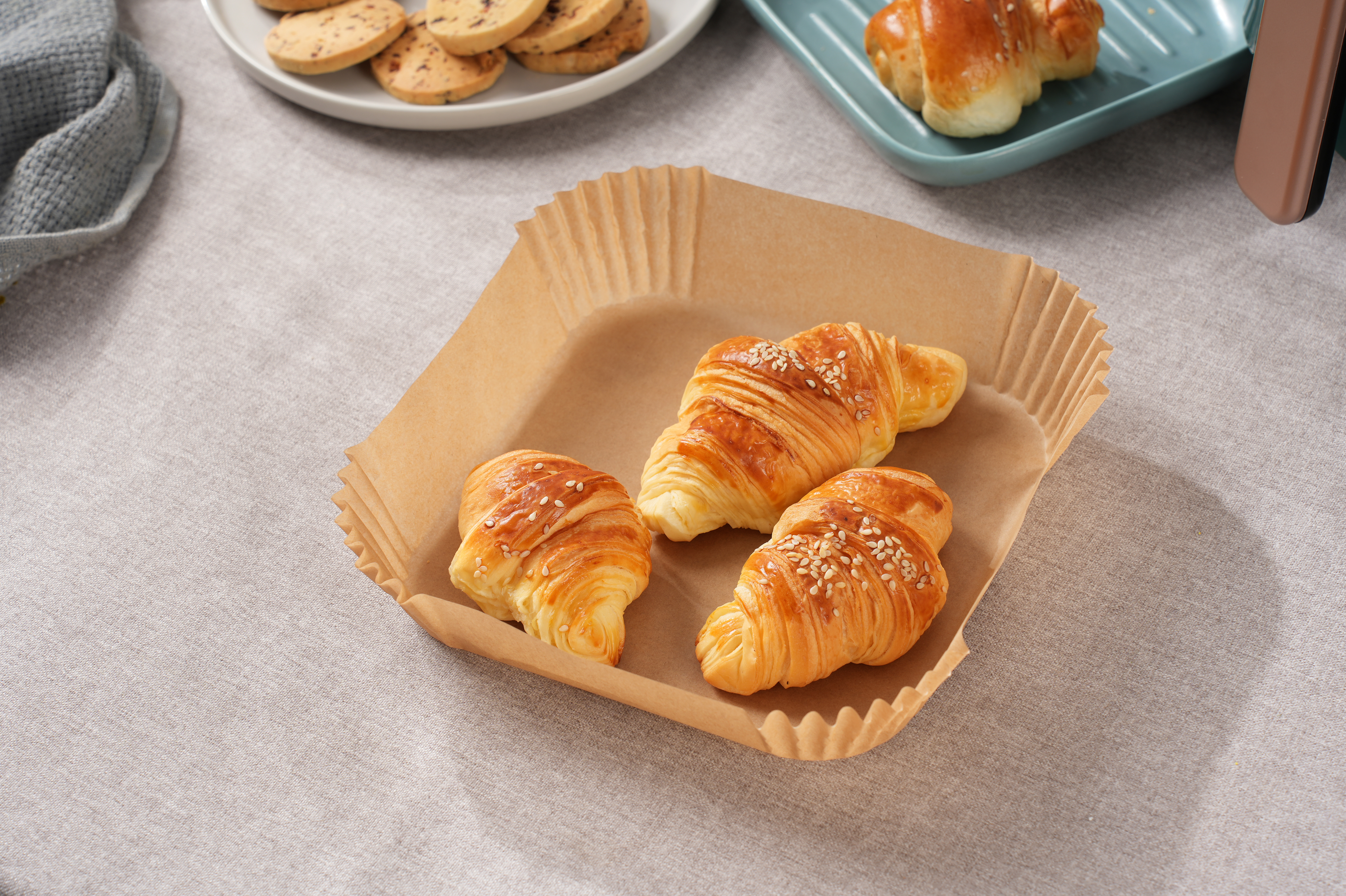 Trzy rogaliki w papierowej tacce na stole z ciastkami i niebieskim talerzem.
