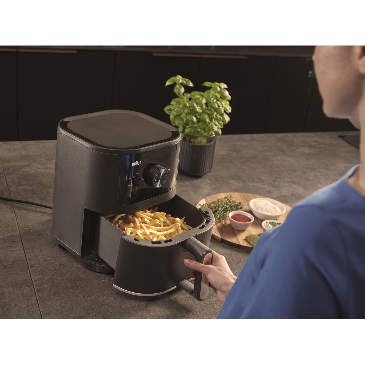 Zwarte airfryer met friet. Vrouw houdt de handgreep vast. Keukenomgeving met kruiden en specerijen.