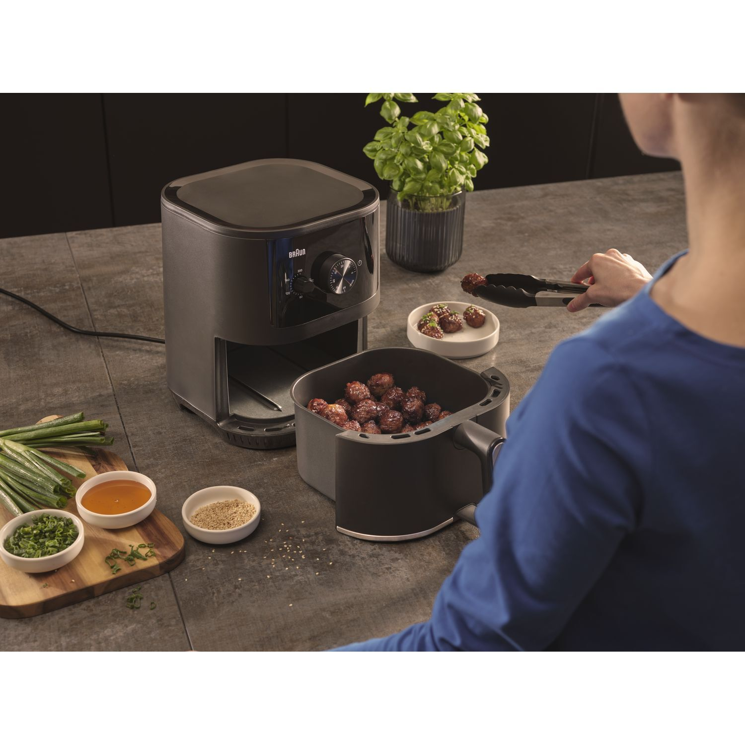 Een airfryer met gehaktballen, saus en kruiden. Een persoon gebruikt een tang.