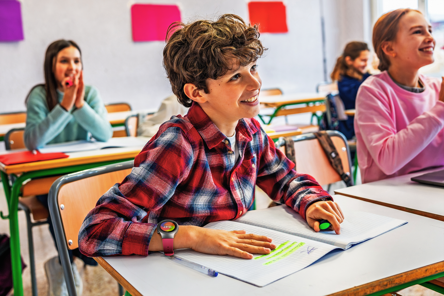 Un niño en un aula sonríe, usando un reloj inteligente. Otros en el aula.