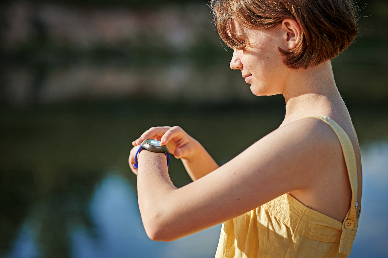 Una chica mira su reloj inteligente junto al agua. Lleva un top amarillo, reloj con correa azul.
