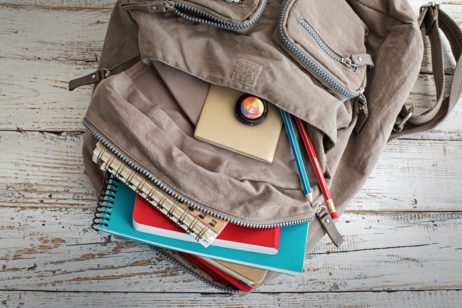 Una mochila está abierta, con un cuaderno, lápices y un reloj inteligente dentro, sobre una mesa de madera blanca.