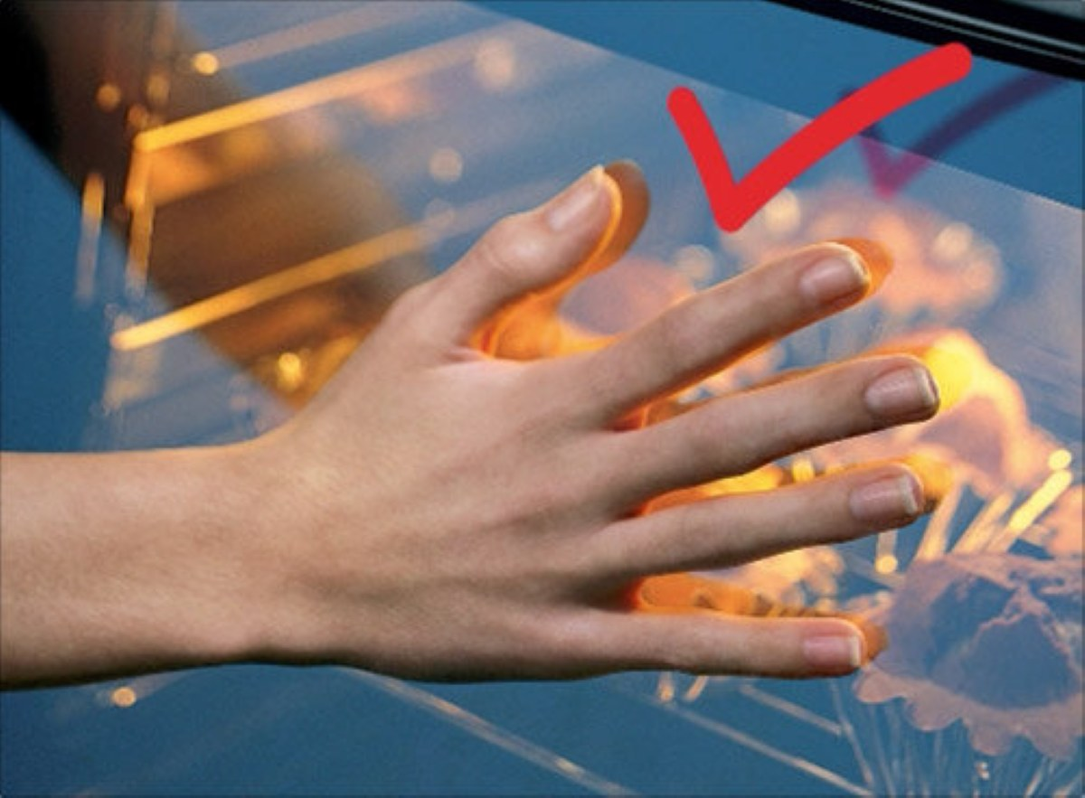 Dłoń dotykająca szkła piekarnika z czerwonym znakiem wyboru wskazującym bezpieczeństwo.