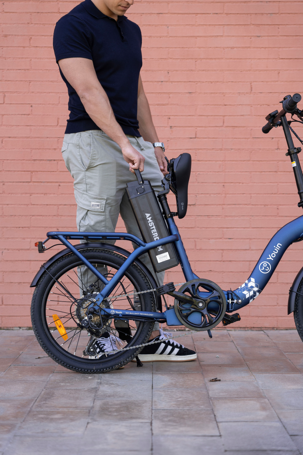 Un hombre sosteniendo una batería de una bicicleta eléctrica azul. Está parado en una pared de ladrillos.