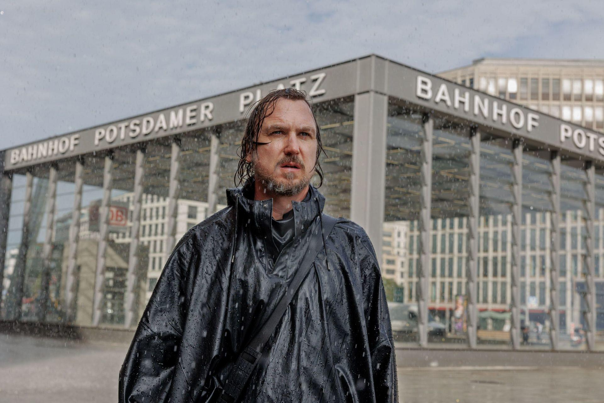 Mann in schwarzem Regenmantel vor einem Gebäude mit der Aufschrift 'Bahnhof Potsdamer Platz'.