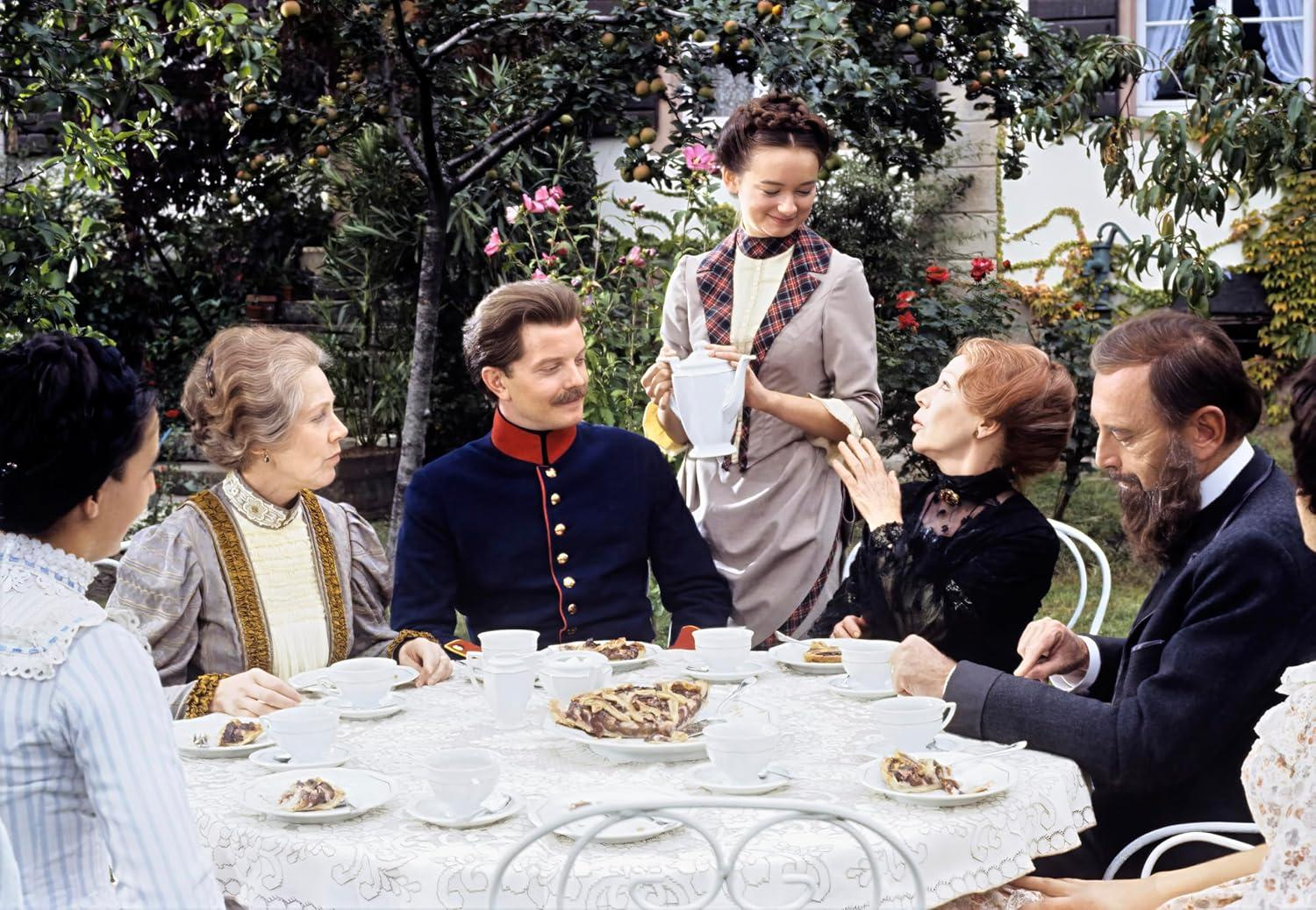 Eine Gruppe von Leuten sitzt in einem Garten an einem Tisch. Eine Frau gießt Tee ein.