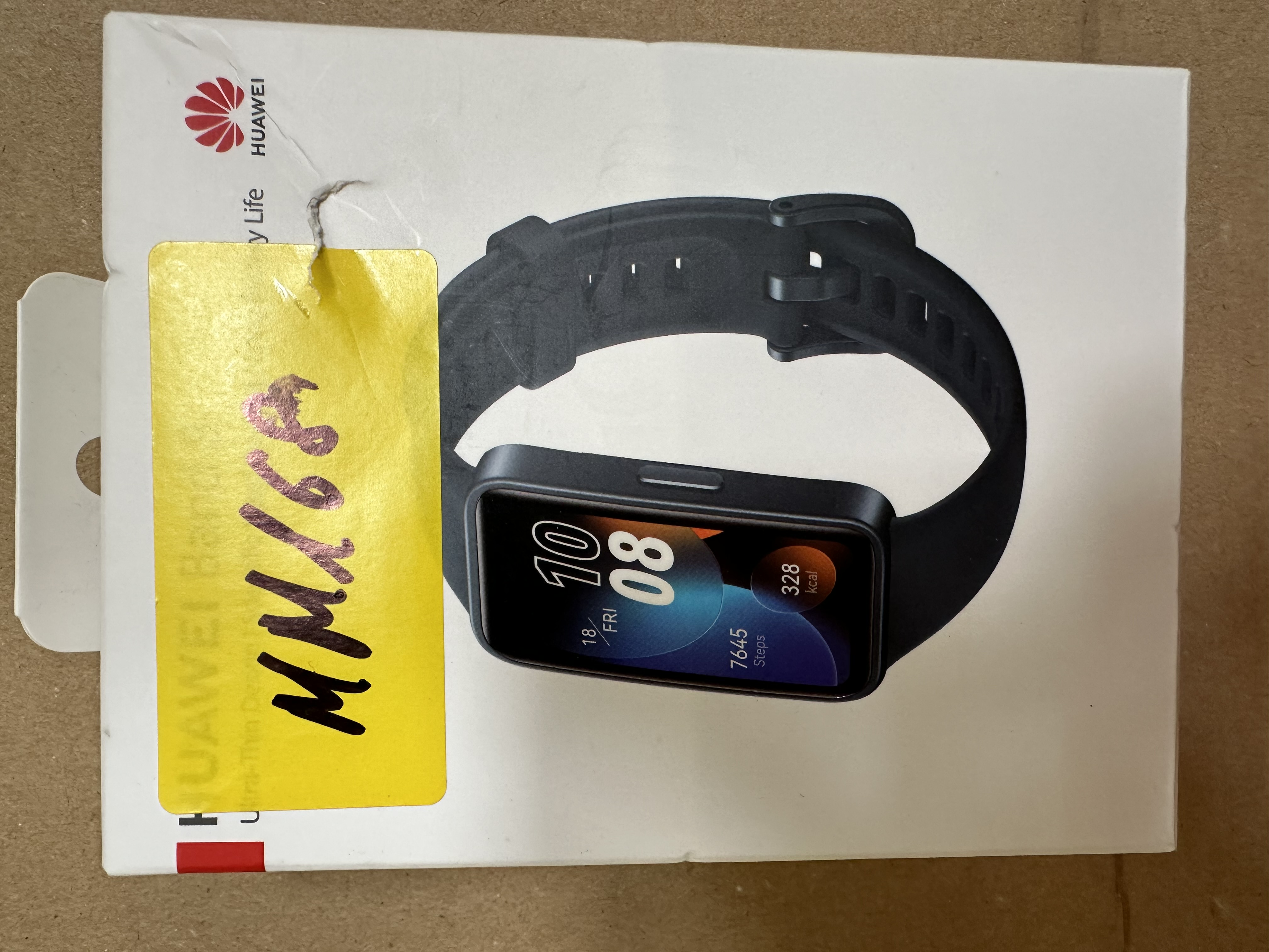 Pudełko z czarnym smartwatchem Huawei. Zegarek wyświetla godzinę, datę, kroki i kalorie. Dołączona żółta etykieta.