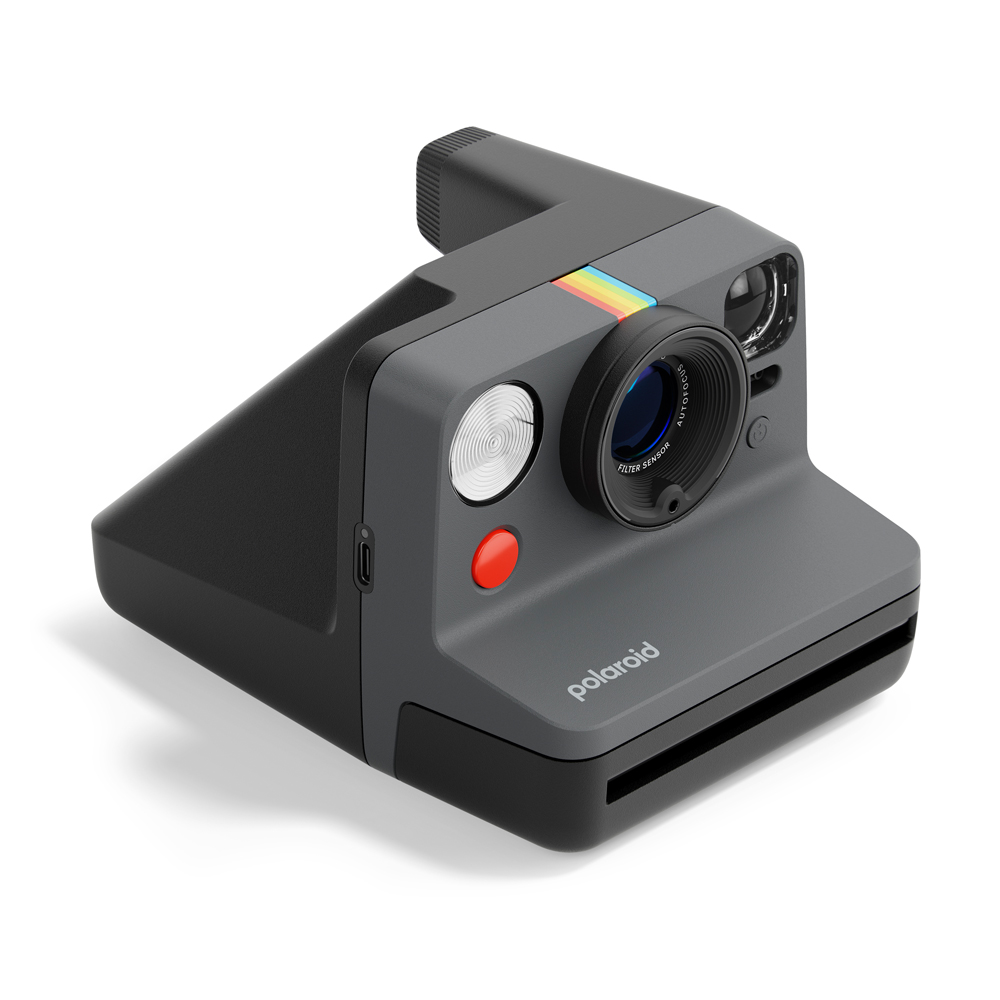 Merceğinde gökkuşağı şeridi olan koyu gri bir polaroid kamera. Kamera beyaz bir yüzeyde.