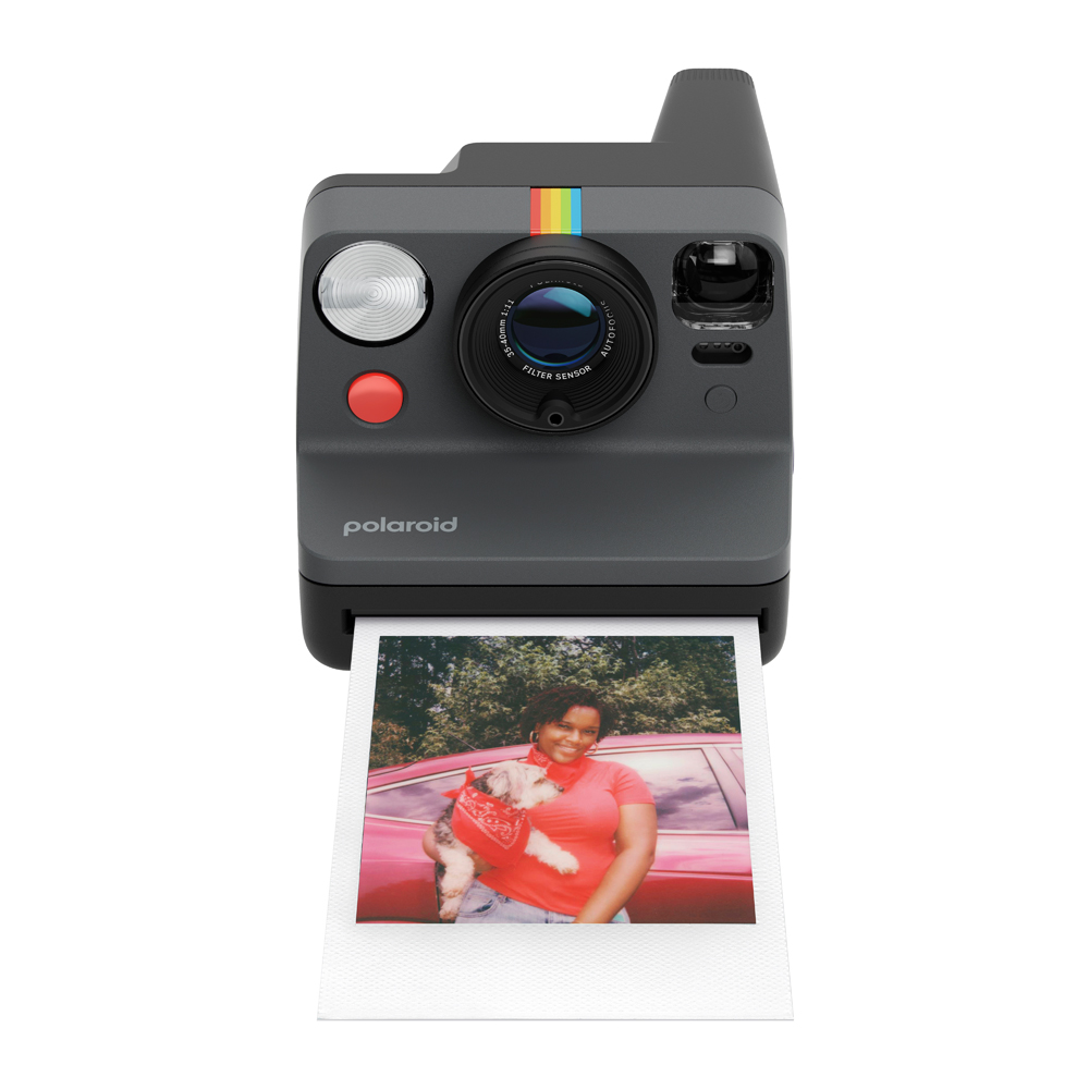 Polaroid kamera, bir araba önünde köpek tutan bir kadının fotoğrafını basıyor.