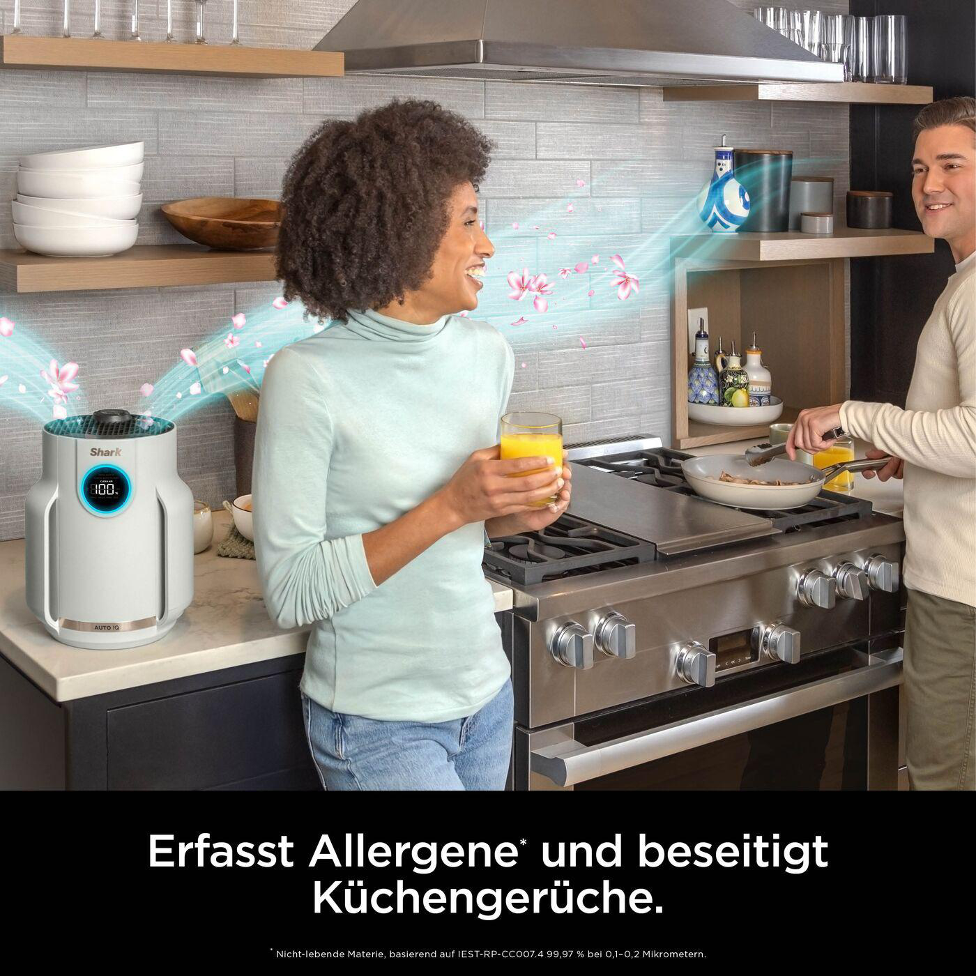 Paar in der Küche, Luftreiniger in der Nähe. Frau mit Getränk, Mann kocht. Saubere Luft wird mit Blumen visualisiert.