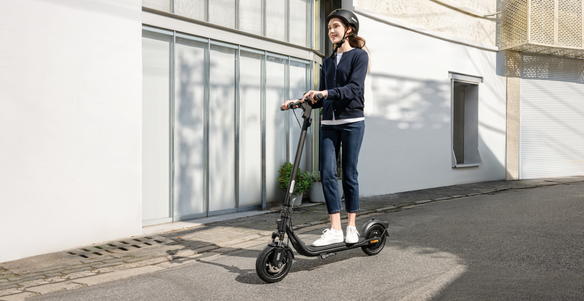 Femme conduisant une trottinette électrique noire dans la rue. Elle porte un casque et une veste bleu foncé.