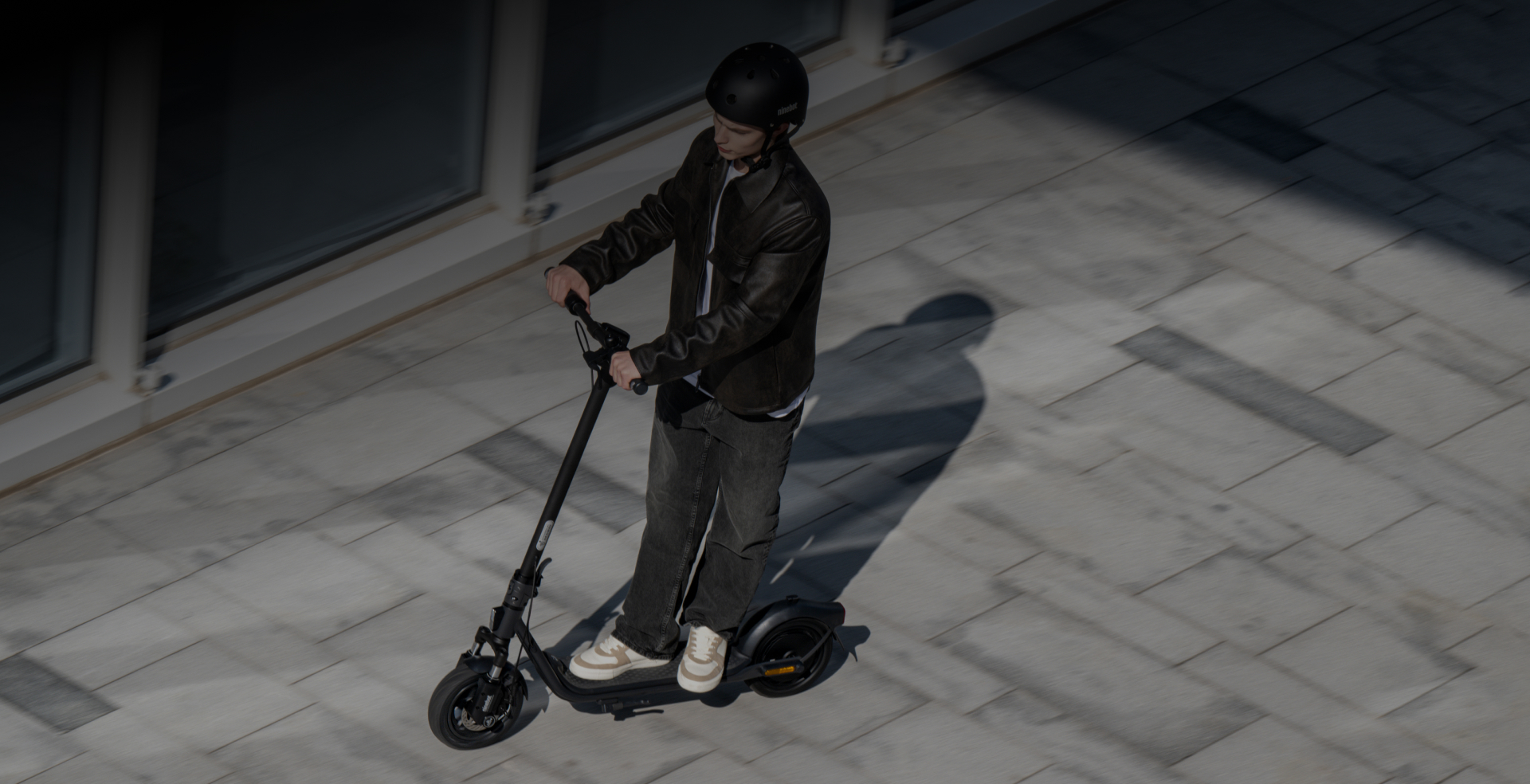 Homme sur une trottinette électrique noire roulant sur une allée. Il porte une veste noire et un casque.