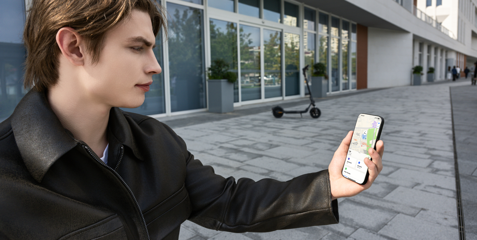 Homme tenant un téléphone avec une carte, avec une trottinette électrique et un bâtiment en arrière-plan.