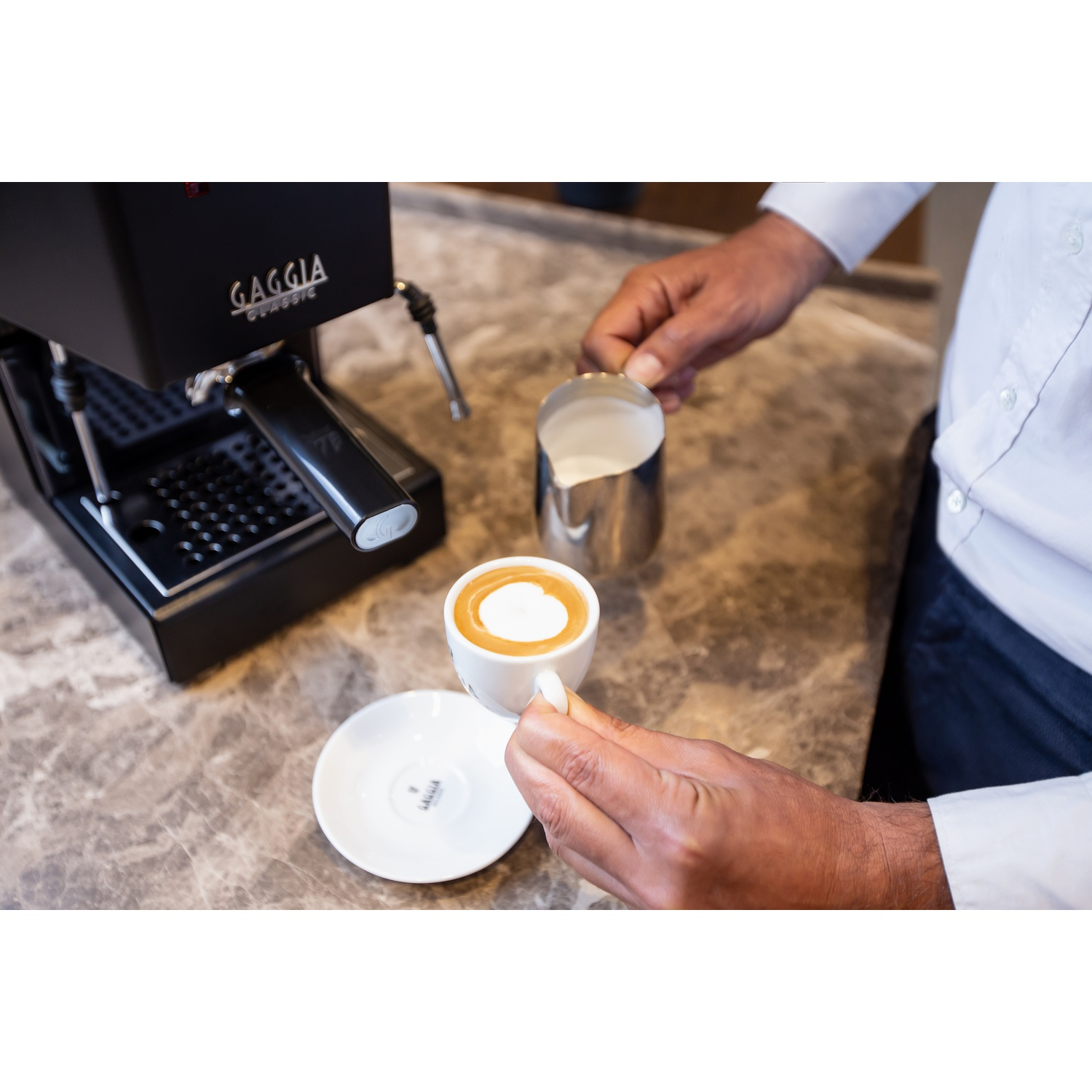 Una persona tiene in mano una tazza di espresso e un bricco di latte vicino a una macchina per il caffè.