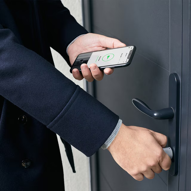 Persona desbloqueando una puerta con su teléfono, manija visible, colores neutros.