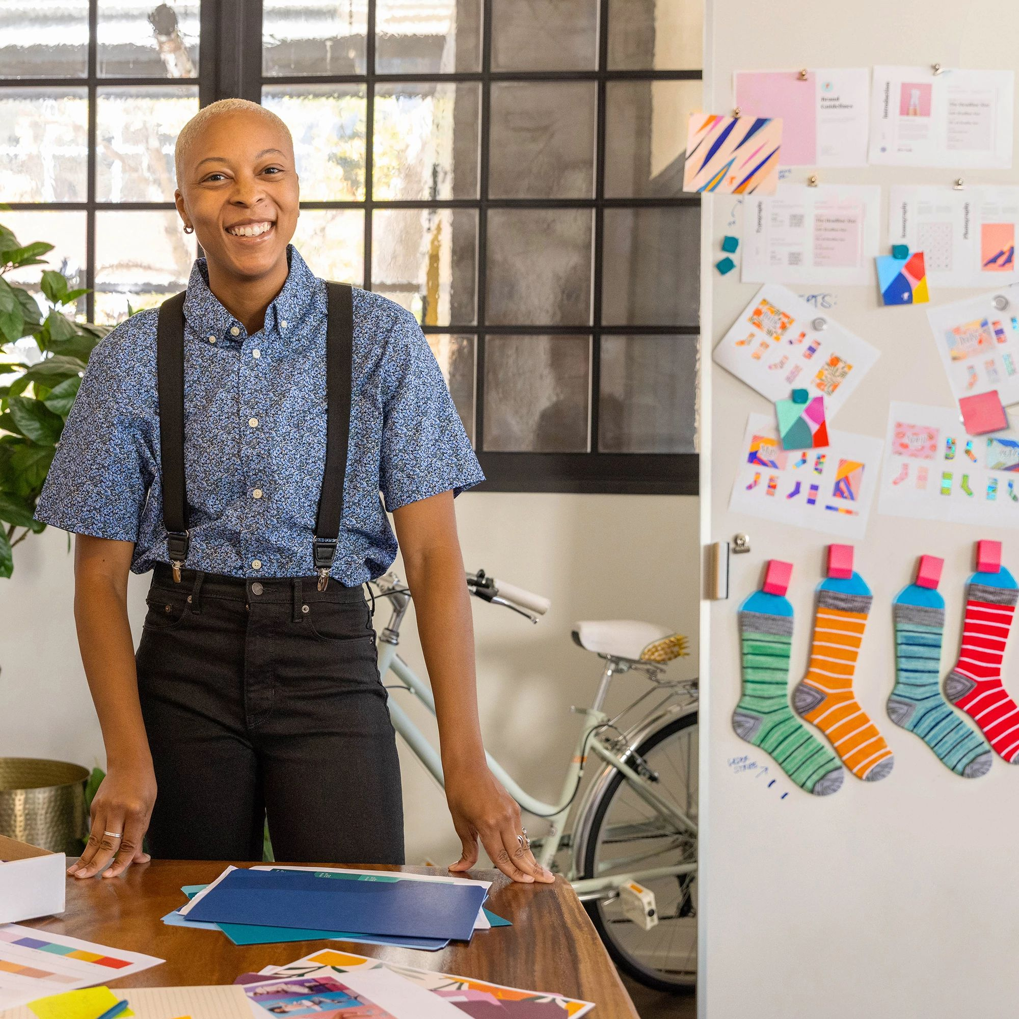 A woman is smiling. She is in an office with a desk and a window. There is a bicycle and a fridge with socks.