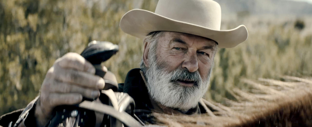 Alec Baldwin in einem Cowboyhut, lächelnd und hält einen Sattel.