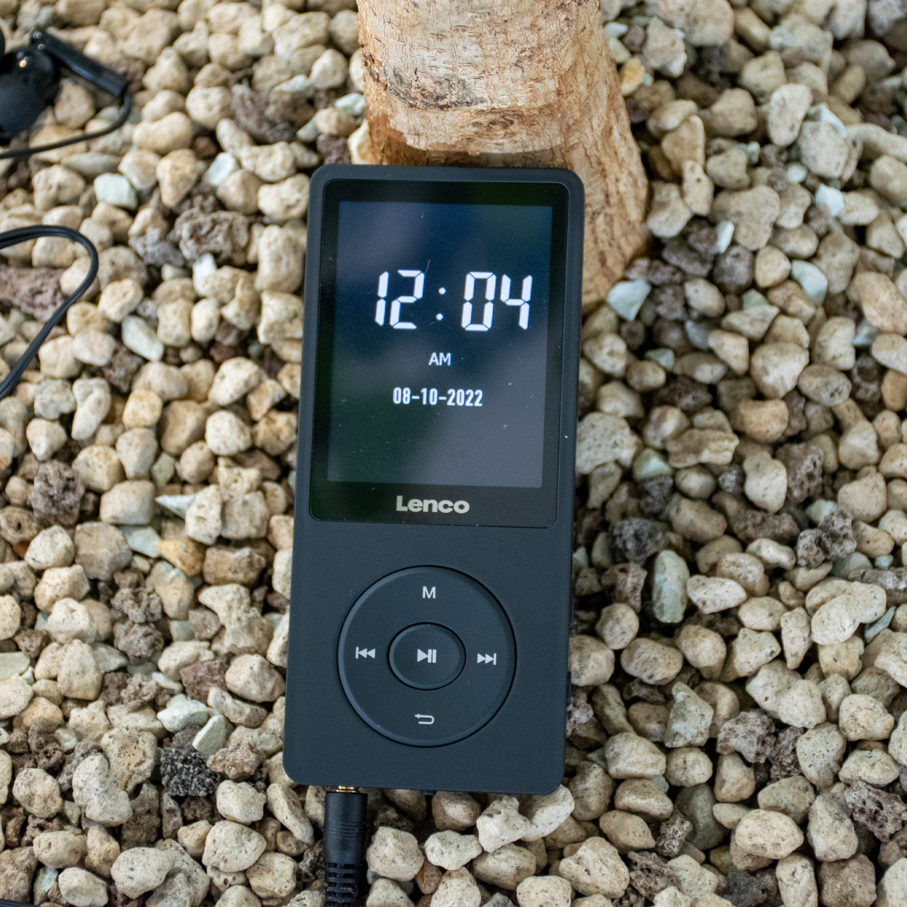 Lettore MP3 nero con display dell'ora, su un letto di ciottoli, con un tronco d'albero sullo sfondo.