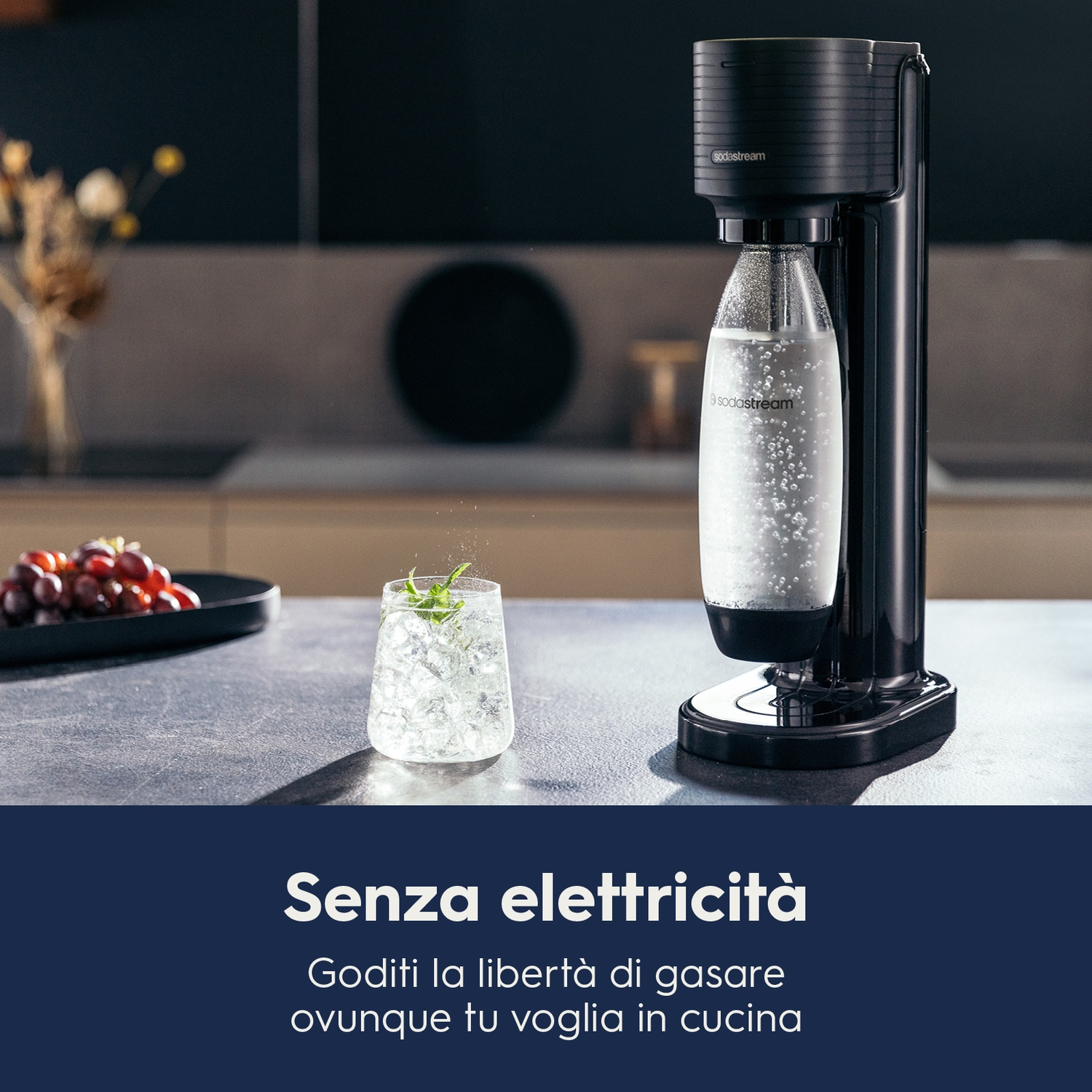 Una macchina SodaStream nera con un bicchiere d'acqua e sfondo cucina. Uva a sinistra.