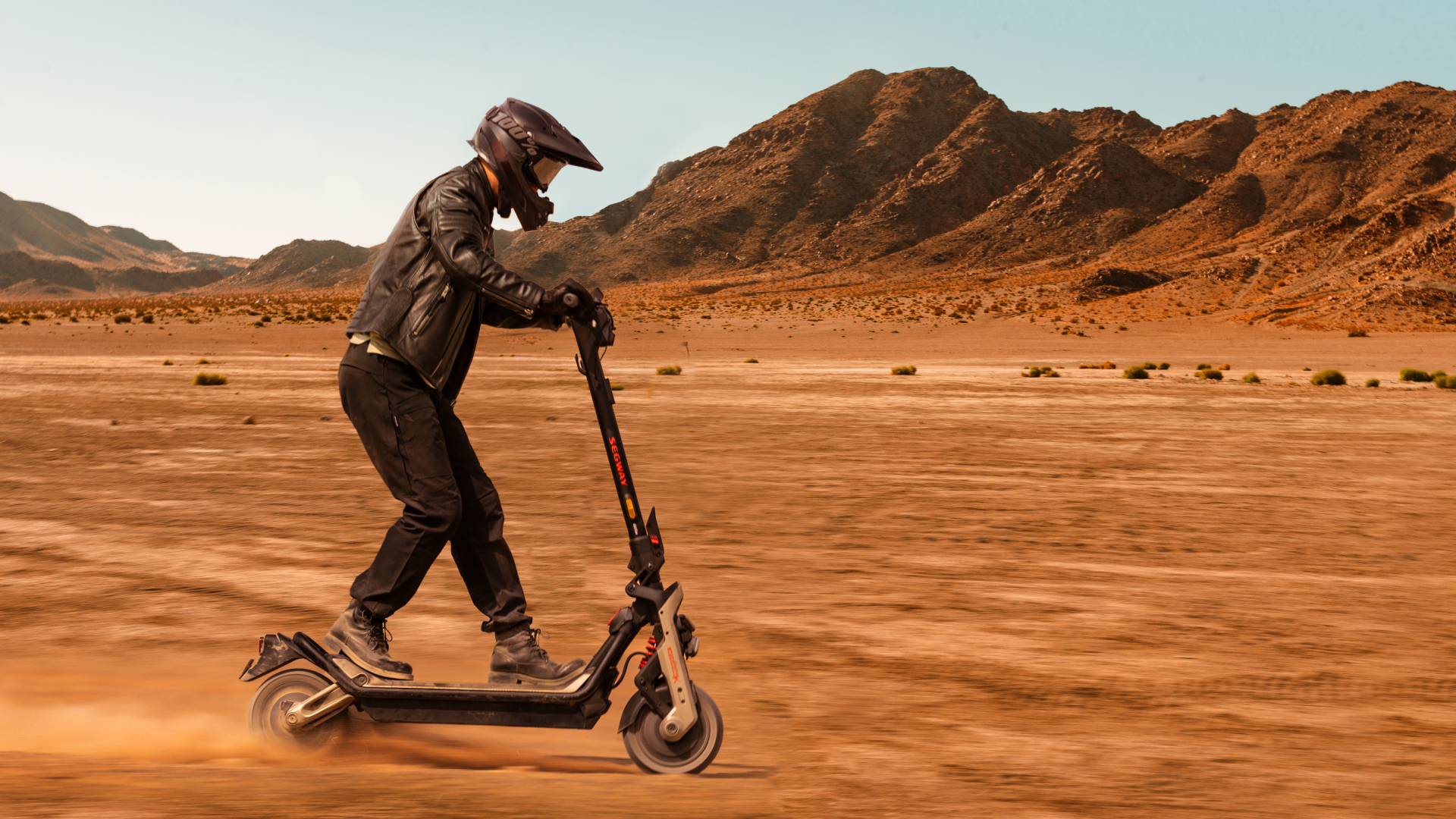 Homme conduisant un scooter électrique dans un désert, portant un casque et une veste en cuir.